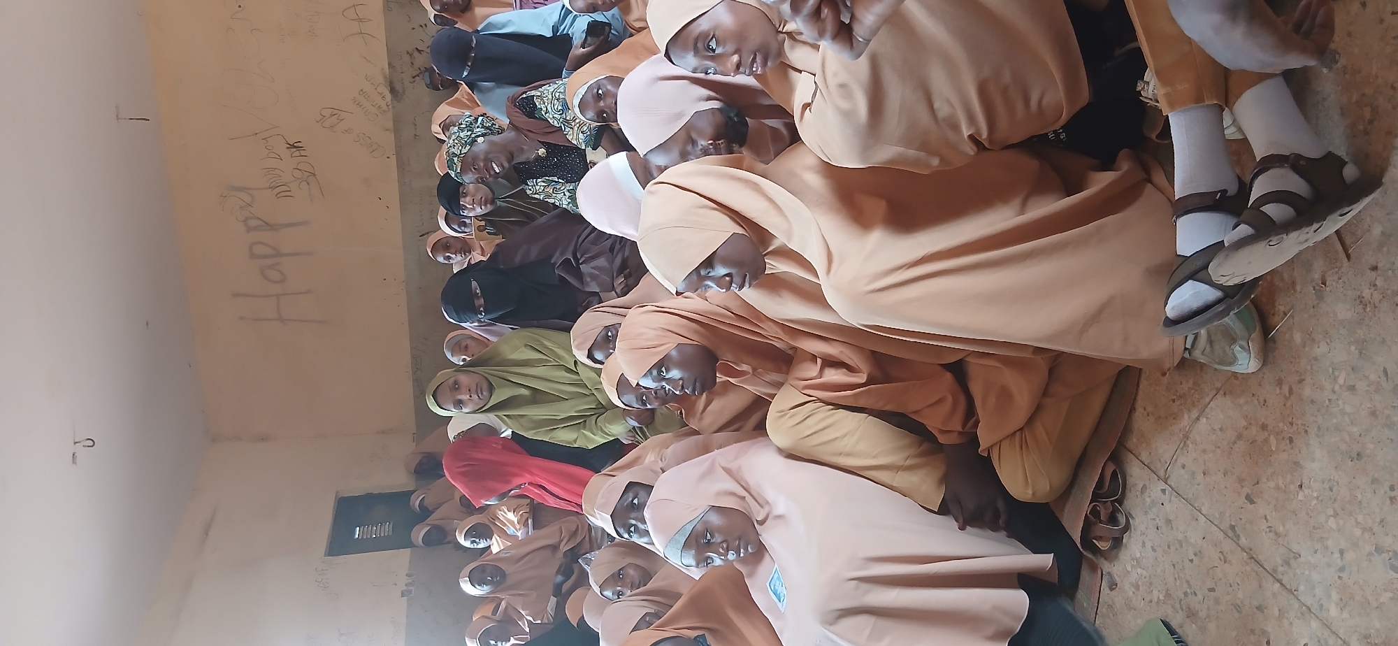The image depicts an indoor scene at a school in Hayin Banki, Nigeria. A group of young women, likely students, are seated on the floor, facing away from the camera and towards the front of a classroom. They are all wearing modest clothing, with most of them wearing headscarves or hijabs in various shades of light brown and beige. Some are wearing black niqabs, covering their faces.
The environment appears to be a classroom with a light-colored, possibly concrete floor. The walls are painted a light, neutral color, and are covered in graffiti. Words and symbols are scrawled across the upper portion of the wall, including what appears to be "Happy" written in large letters. A metal hook is visible on the ceiling. The lighting in the room is somewhat dim, suggesting it might be an overcast day or the room relies on natural light from an unseen source.
In the foreground, the feet and lower legs of some of the individuals are visible. They are wearing sandals and some have socks. The overall impression is of a gathering of students in a classroom setting, possibly attending a lesson or an event. The presence of graffiti on the walls suggests a well-used and perhaps aged educational facility.