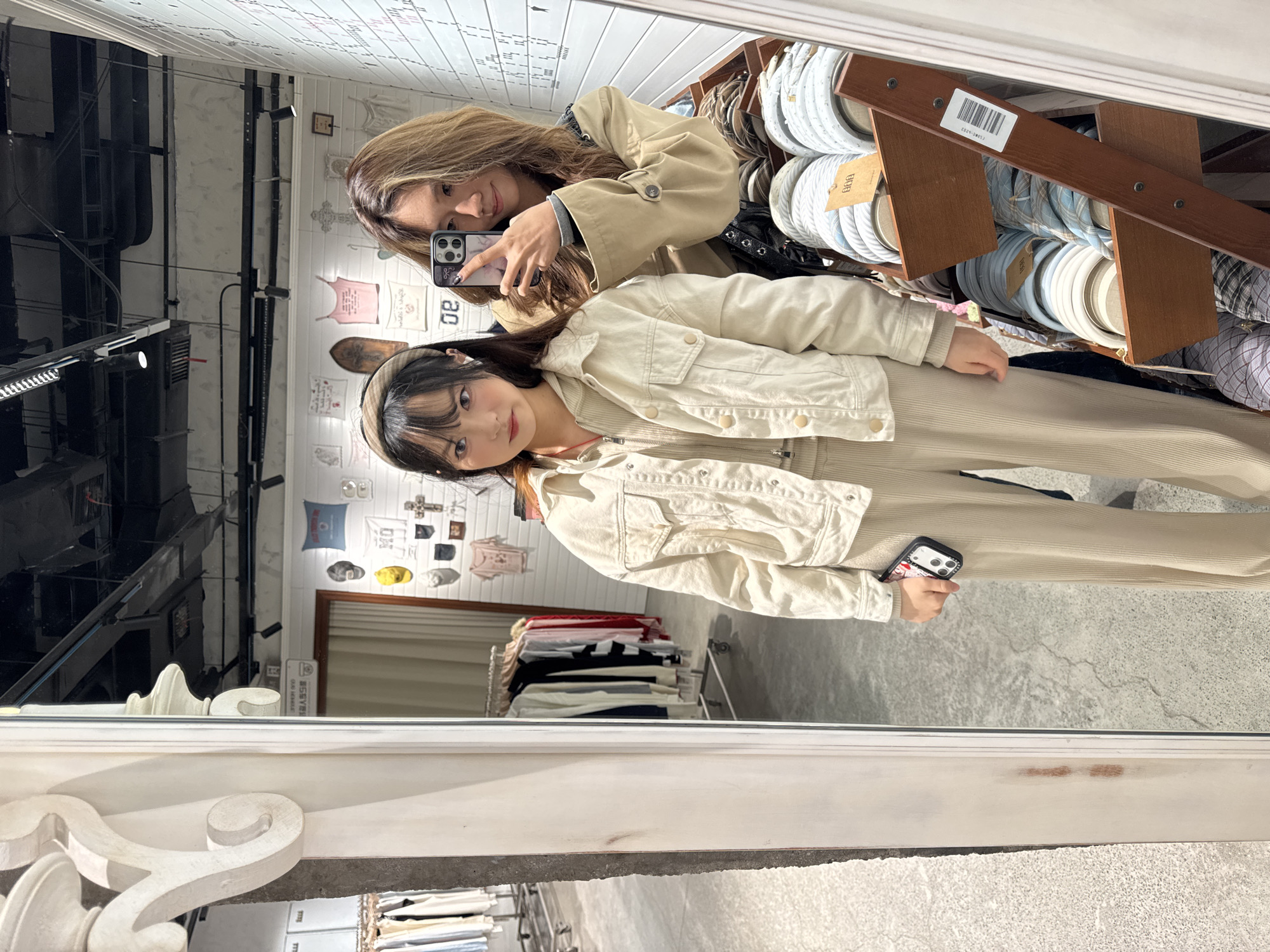 Two women are taking a selfie in a mirror in a shop in Jianlan Road District, China. The women are wearing light-colored clothing. The woman on the bottom is wearing a headband. Shelves with stacked rolls of fabric are to the right of the women. Various clothing racks with different items are reflected in the background. The mirror has an ornate, decorative white frame.