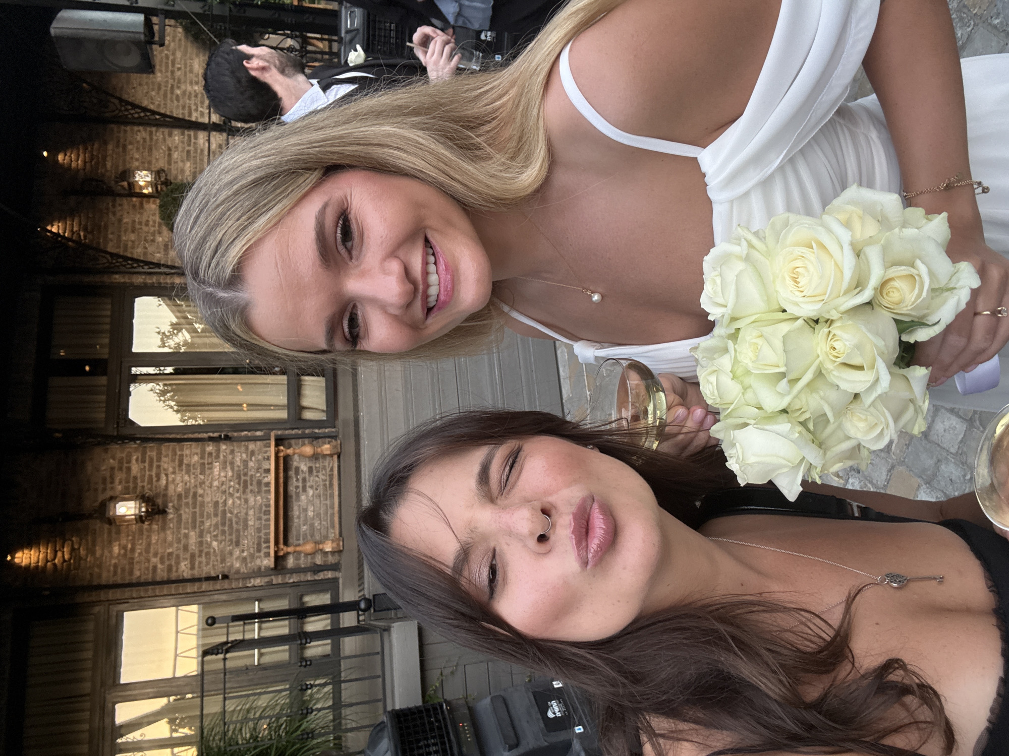 Two women are posing for a selfie in what appears to be an outdoor venue. The woman on the left has fair skin, blonde hair, and is wearing a white dress. She is holding a bouquet of white roses and smiling at the camera. The woman on the right has tan skin, brown hair, and is wearing a black top. She is pouting and winking at the camera.
The background shows an outdoor setting with a brick building featuring windows and decorative shutters. Metal railings and outdoor lighting fixtures are also visible. A speaker and other equipment are in the background. People in formal attire are seen in the background, suggesting the event is a wedding or formal party.
The location can’t be determined as Tel Aviv as no specific buildings and landmarks are discernible in the image.