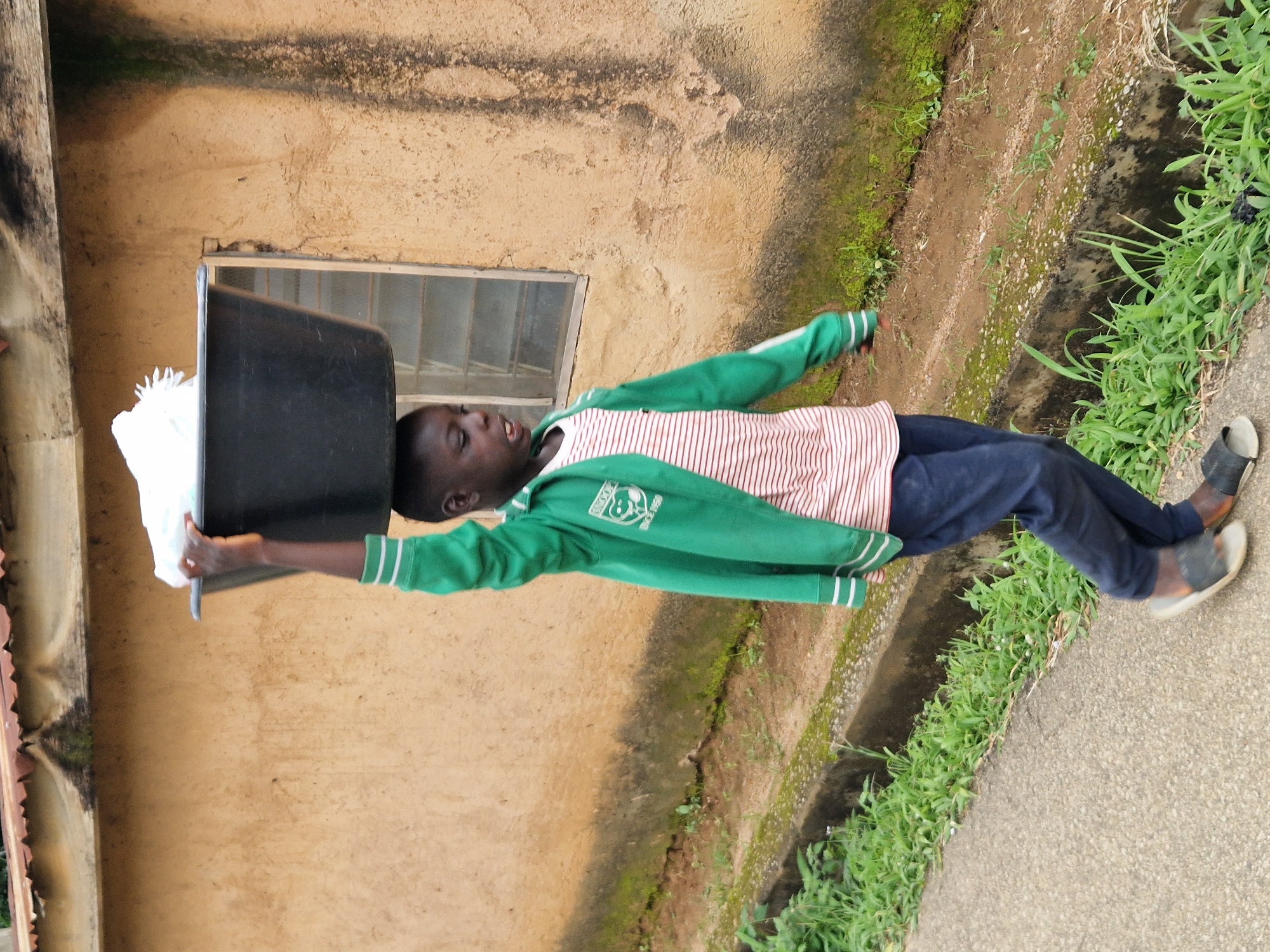 A young Black boy is walking on a paved path in Jos, Nigeria, carrying a large black tub filled with white items on his head. He is wearing a green hooded jacket with white stripes on the sleeves and a red and white striped t-shirt underneath. He also has on dark blue pants and dark sandals. The boy is looking down and to his right, with his mouth open. The background features a tan stucco building with a window and a rusty metal roof. The ground beside the path is covered with green grass and a concrete drainage ditch. The scene appears to be a residential area in Jos, Nigeria.