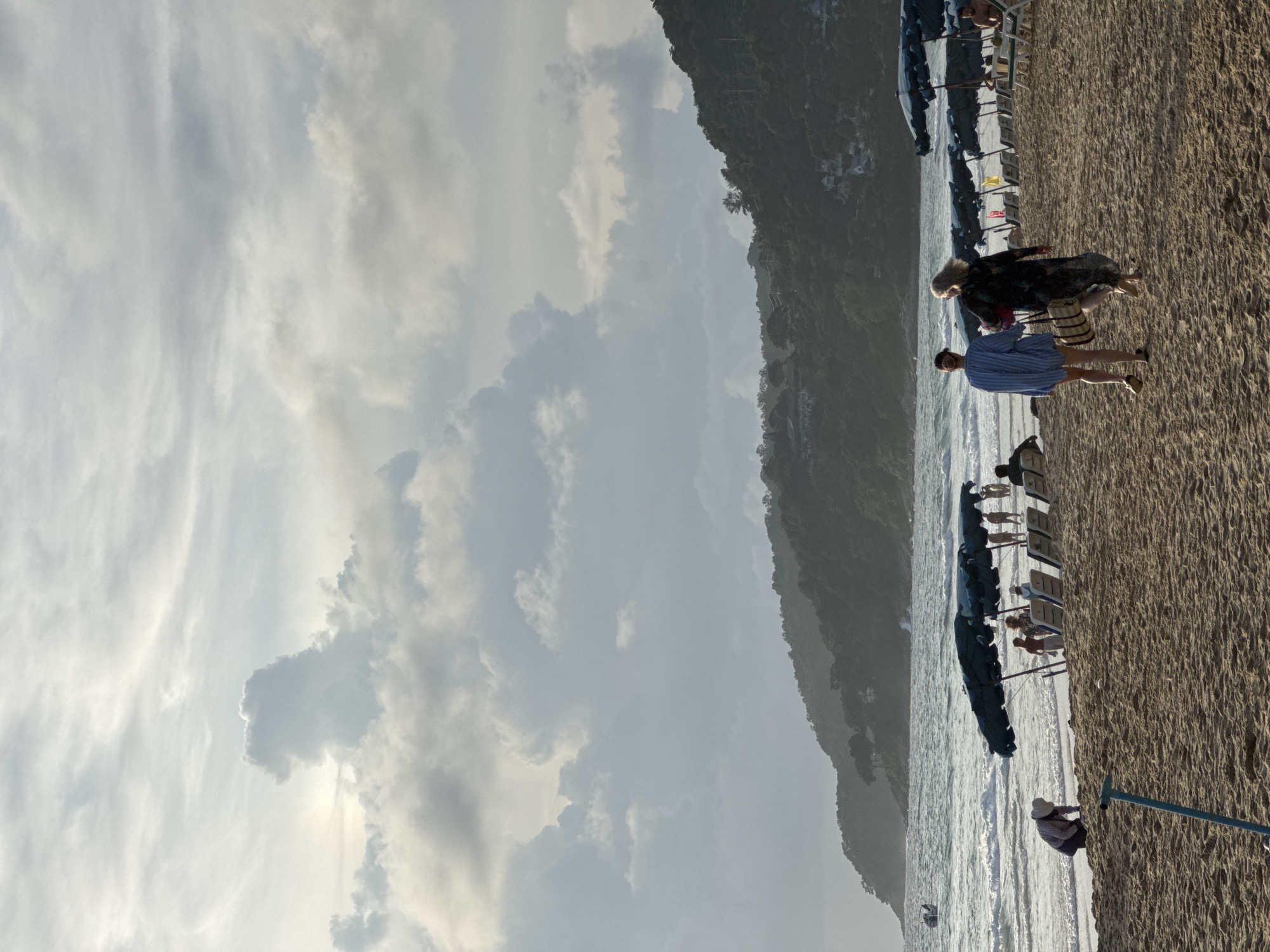 On a sandy beach in Karon, Thailand, two individuals walk away from the camera towards the water. The person on the left is wearing a blue and white striped oversized shirt and light-colored shorts. The person on the right is wearing a long, dark patterned dress and carrying a straw bag. They are walking on the beach, which is lined with rows of lounge chairs and umbrellas. In the background, the ocean is visible with waves and a distant boat. The horizon is marked by a lush, green, tree-covered hill. The sky is overcast with dramatic, textured clouds. Closer to the foreground, a person in a hat is bent over on the sand.