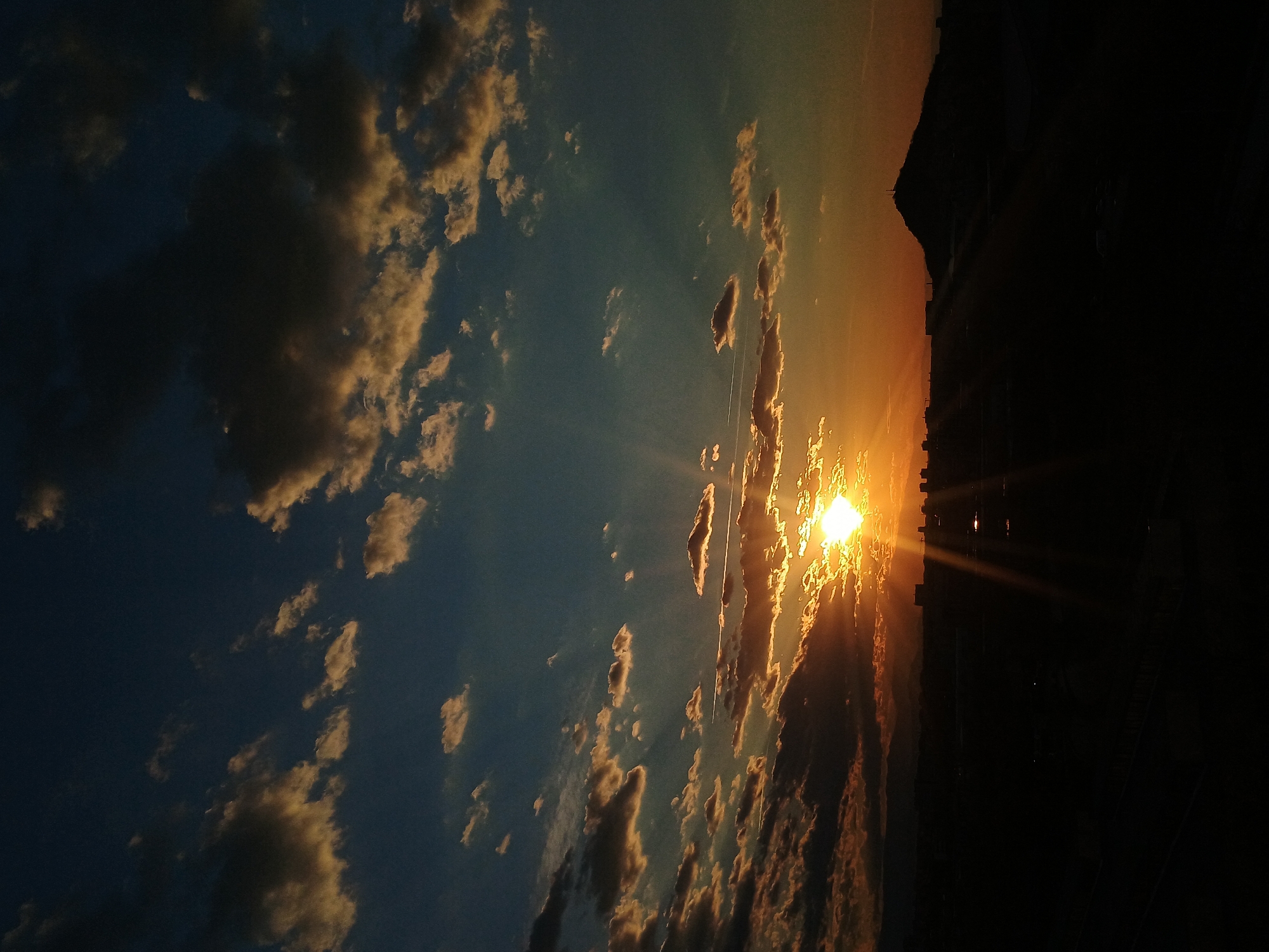 The image shows a sunset in Plovdiv, Bulgaria. The sun is low on the horizon, shining brightly and creating rays of light. There are numerous clouds in the sky, with some silhouetted against the light and others illuminated with golden hues. The silhouette of buildings and possibly a hill or mountain is visible on the right. The city portion is mostly in deep shadow, with a few points of light.