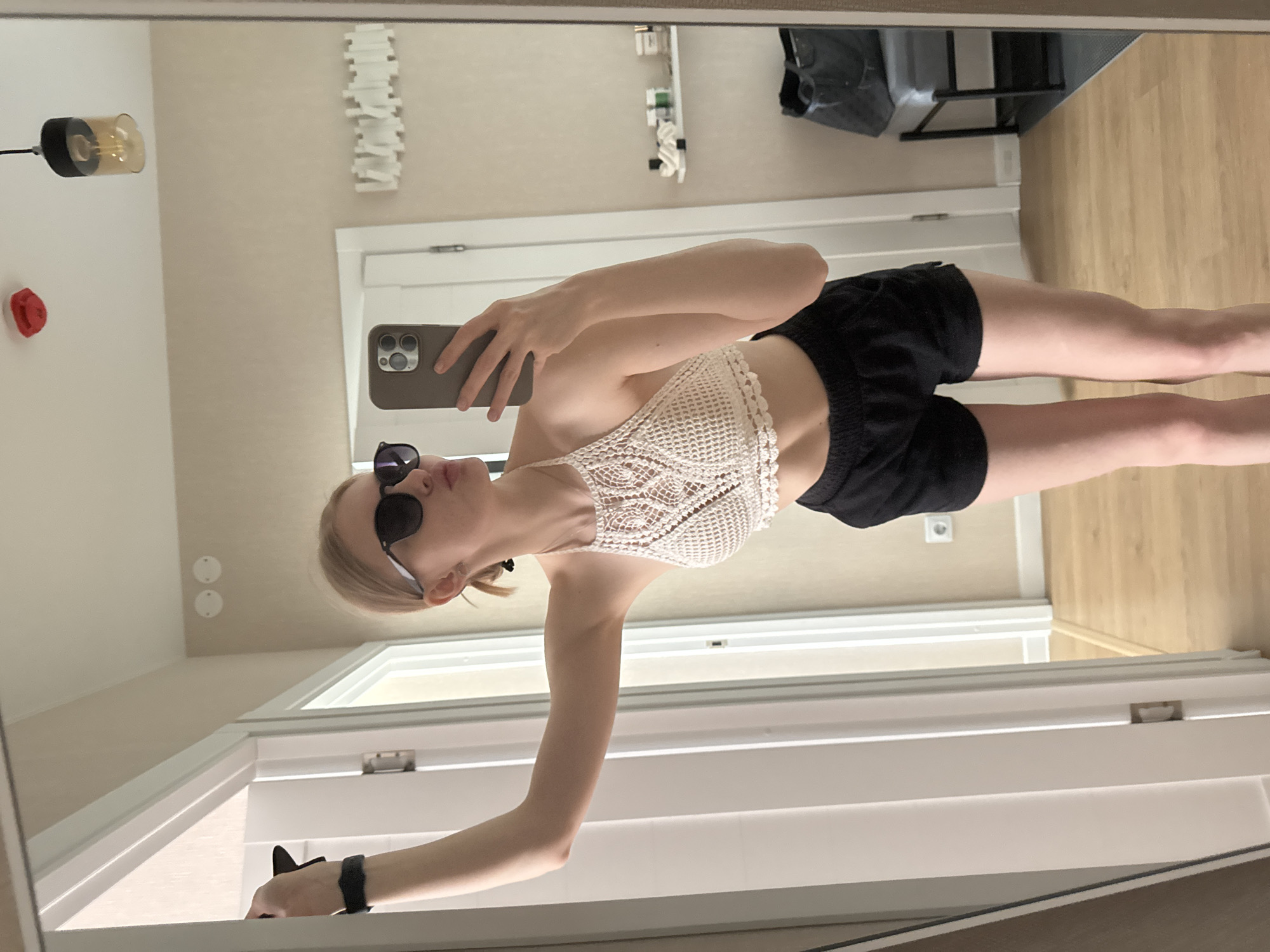 A young woman is taking a selfie in a mirror. She is wearing sunglasses, a white crocheted halter top, and black shorts. The background includes a light fixture, a door, a piece of decorative art, and a bag on a chair. The location is Perm, Russia.