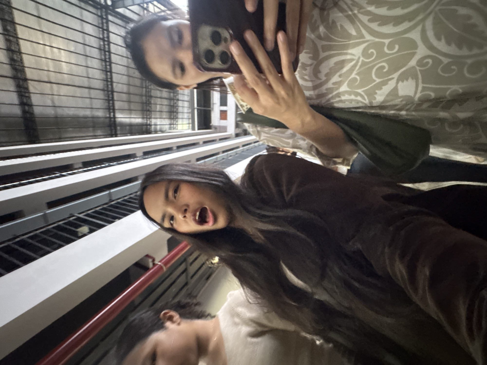Three individuals are captured in a selfie-style photograph, likely taken in a covered walkway or transport terminal in Davao City, Philippines, judging by the architectural style with metal roofing and support structures visible in the background.
In the foreground, a young woman with long, dark hair is looking towards the camera with her mouth open in an exaggerated expression. She is wearing a dark brown jacket over a lighter colored garment. To her right, another young woman is holding a smartphone in her right hand, seemingly looking at it. She is wearing a light-colored, patterned top and has a dark green bag slung over her shoulder. Partially visible on the far left is a third individual, also appearing to be a young woman, wearing a light-colored sweater, with her head turned away from the camera. The overall lighting suggests it might be daytime, possibly indoors or in a shaded outdoor area.