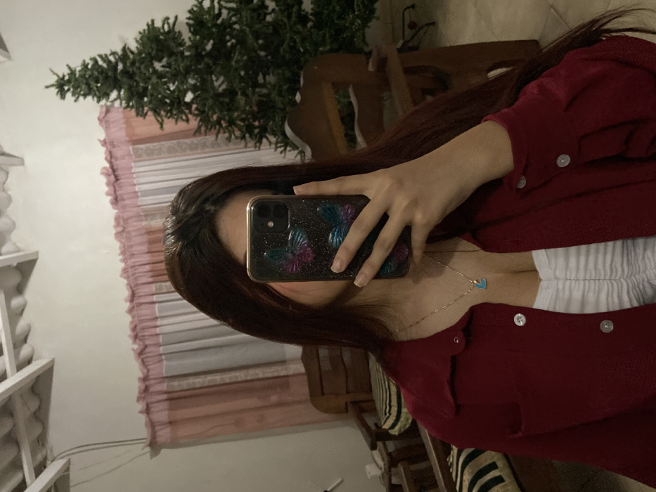 A person stands indoors, taking a selfie in a mirror. They wear a red button-down shirt over a white top. A gold necklace with a blue heart pendant is visible. The phone has a butterfly case. Behind them, a blurred scene reveals a green Christmas tree, a pink curtain, and a wooden chair. The photo is rotated to the left.