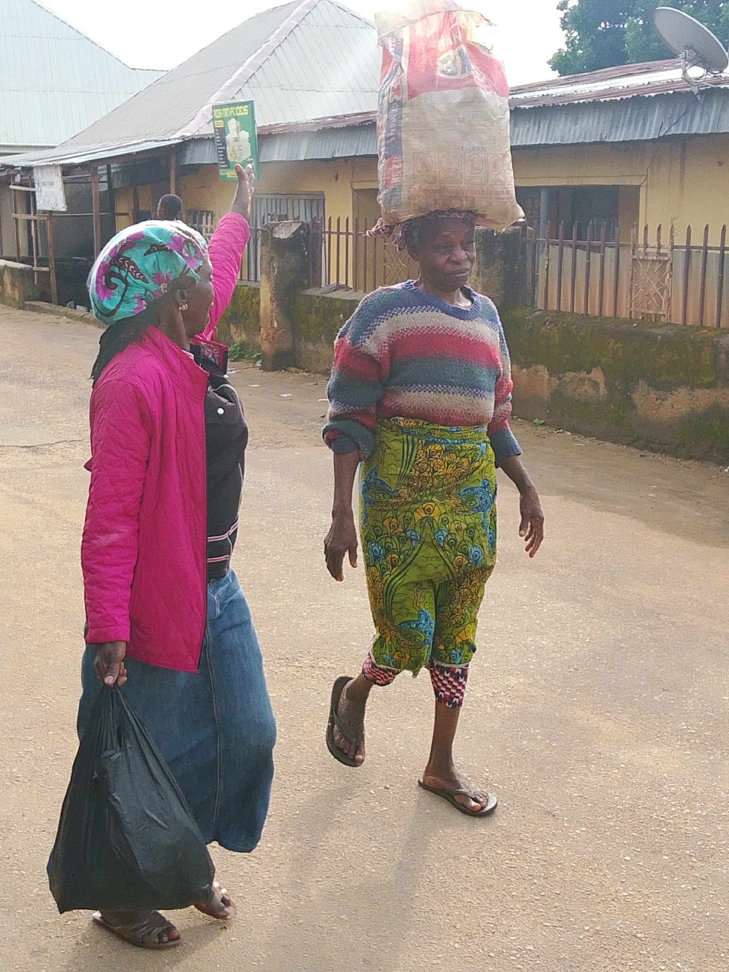 Two women are walking on a paved road in Jos, Nigeria. The woman on the left is wearing a pink jacket, a blue skirt, and a patterned headscarf. She is carrying a black plastic bag in her left hand and holding a green pamphlet in her right hand. The woman on the right is carrying a large sack of goods on her head. She is wearing a striped sweater and a patterned skirt. The environment appears to be a residential or commercial area with buildings and fences in the background. The sun is casting shadows on the road, indicating it is likely daytime.