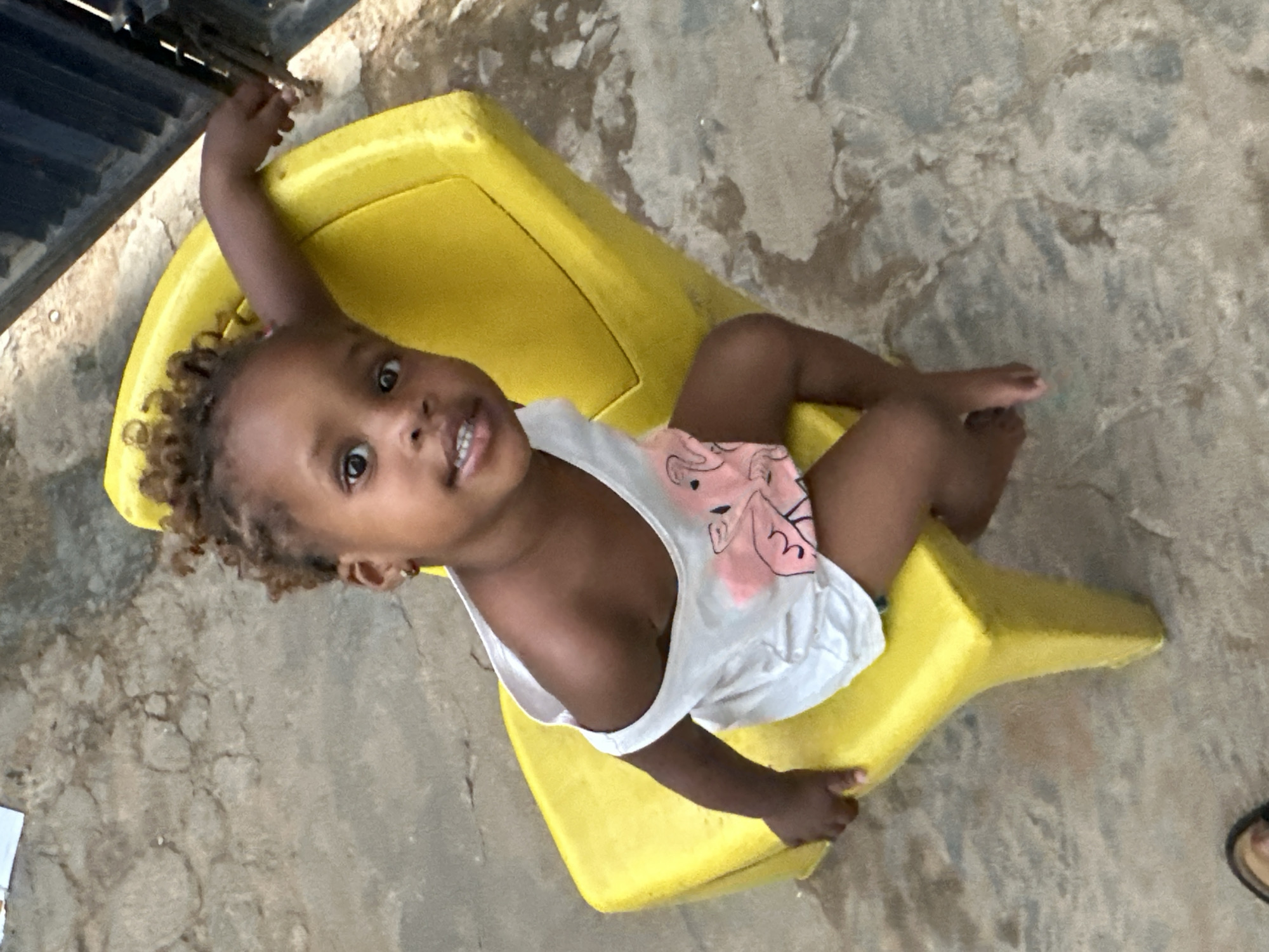 A high-angle, vertical photograph shows a young child sitting on a bright yellow, molded plastic seat or toy. The child, appearing to be a toddler, has dark skin, curly brown hair, and is dressed in a white sleeveless top featuring a light pink design and pink shorts. They look directly at the camera with wide, brown eyes and a slight, open-mouthed smile, revealing their teeth. One arm is extended upwards towards the top left of the frame, with the hand reaching towards a dark, possibly metallic, structure that suggests a fence or part of a building.
The setting appears to be outdoors or semi-outdoors in Narayi, Nigeria, with the child positioned on a rough, mottled grey-brown ground that resembles concrete or packed dirt, showing various textures and patterns. The scene is well-lit, indicating it is daytime. In the bottom right corner, a small portion of an adult's foot wearing a dark sandal is visible. The child's expression conveys engagement and happiness.