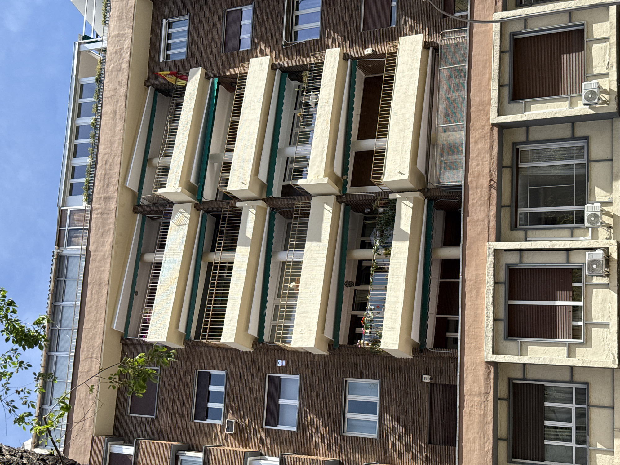 The image shows the exterior of a tall apartment building, photographed from a low angle. The building has a mixed facade, with sections of dark brown, vertical ridged material contrasting with light brown, stucco-like sections. Windows are rectangular, some with dark blinds or shutters partially closed. Several balconies feature distinctive vertical concrete structures with green canvas elements and gold-colored metal railings. The balconies appear tiered or stacked vertically. Small air conditioning units are visible near some windows. A Spanish flag hangs from one of the balconies. Some vegetation is visible on one side of the building. The sky is visible at the top left corner. Based on architectural style, this is likely a building in the city of Madrid, Spain.