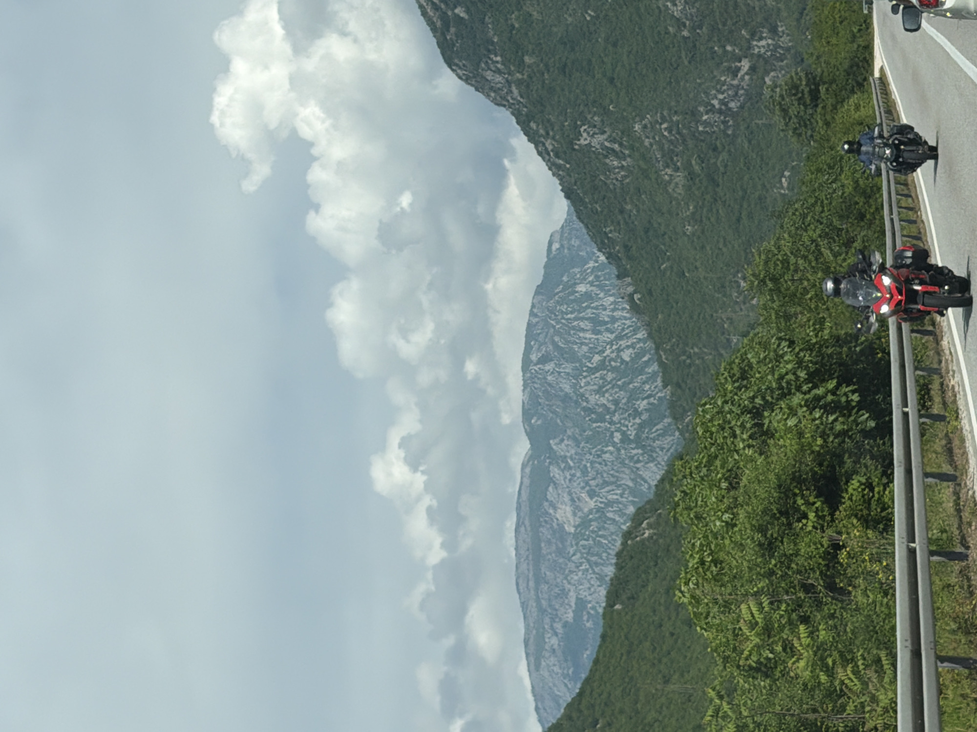 A scenic view shows a road with two motorcycles, a red one in the foreground and a black one further away. The road is bordered by a guardrail and lush green vegetation. A large mountain stretches into the distance, partially obscured by cloudy skies. The location is identified as Donji Jasenjani, Bosnia and Herzegovina.