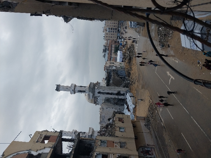 The image presents an elevated view of a street in Khan Yunis, Palestinian Territory, under an overcast sky. The foreground is partially obstructed by dark metal bars, suggesting a vantage point from a damaged building or balcony. The scene depicts widespread structural devastation across the urban landscape.
In the center, a large mosque with a white dome and a tall, slender minaret is prominently visible, exhibiting significant damage to its upper sections and surrounding architecture. To the left, a multi-story building is heavily damaged, with large sections of its facade missing, revealing exposed interiors and structural elements. A poster on this building reads "Gaza Under Attack." Another partially visible damaged building frames the right side of the image.
The street below is a wet asphalt road, marked with a white turn arrow and flanked by extensive piles of rubble, dirt, and debris from collapsed structures. Numerous individuals, including adults and children, are seen walking on the wet road and dirt paths, scattered across the scene. They appear to be civilians moving through the damaged environment. In the mid-ground, further down the street, additional buildings with varying degrees of damage are visible, along with white temporary structures or tents amidst more debris. Vehicles are discernible further in the distance. The overall environment reflects a city heavily impacted by destruction, with its residents navigating the damaged infrastructure.
