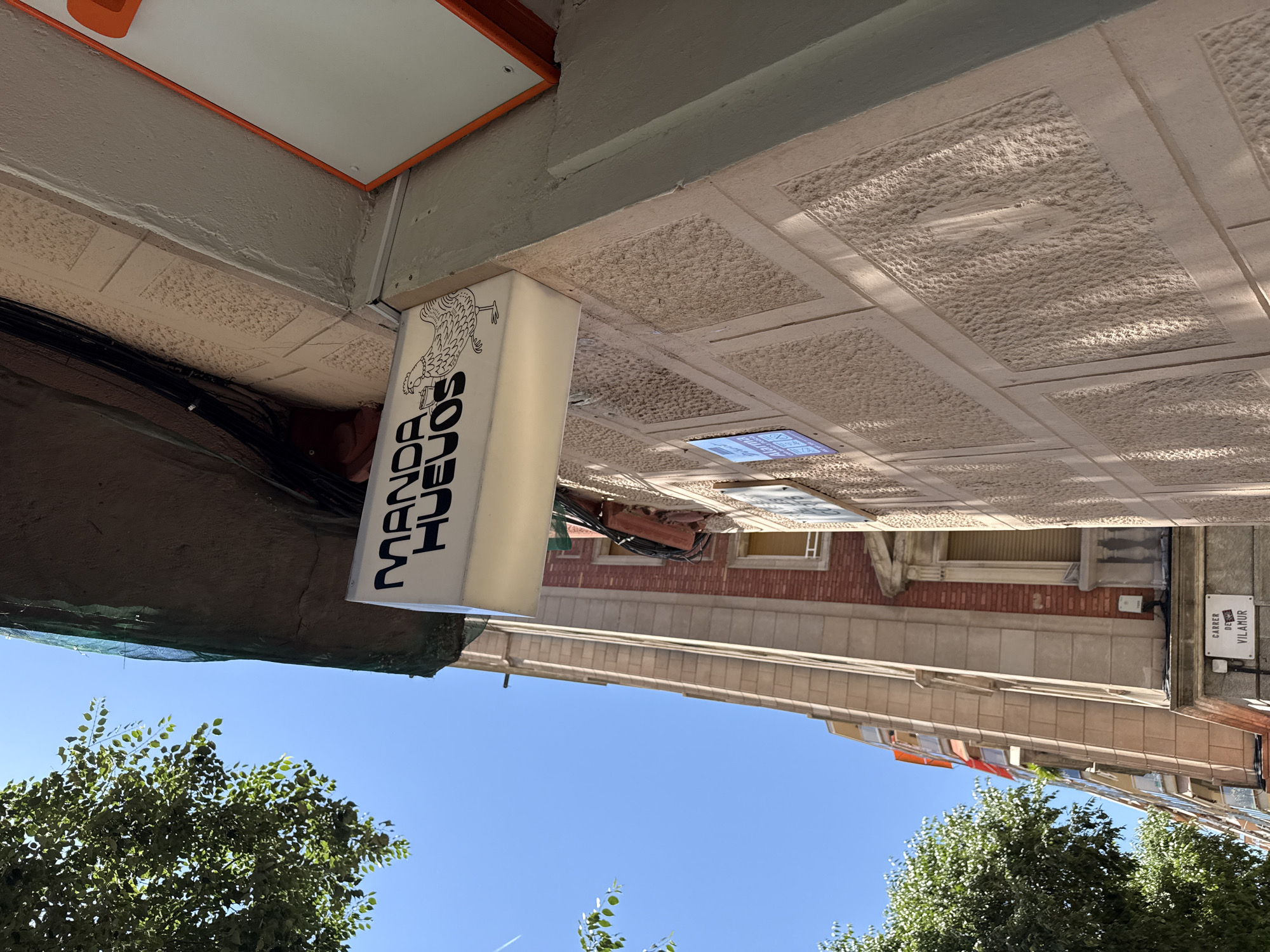 The image depicts a street scene in Barcelona, Spain. The sky is clear and blue, with green treetops in the foreground. The camera is pointing upwards towards the buildings. A sign for "MANOR HUEVOS" is visible, indicating a restaurant. The street sign reads "Carrer de Vilamur". The buildings have brick and concrete facades, and the sidewalk is made of patterned concrete tiles.