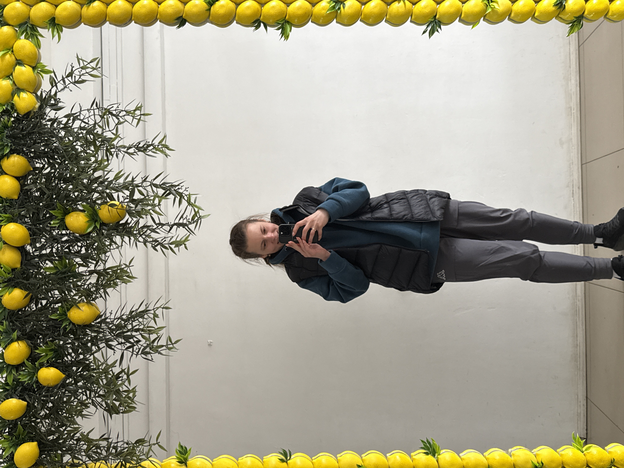 A person stands in front of a wall in what appears to be an interior space. The wall is decorated with an artificial lemon frame and foliage. The person is holding a phone, possibly taking a picture or video. The location is identified as the city of Neftekamsk, Russia. The person is wearing a blue shirt, a black vest, gray pants, and black shoes. The floor appears to be tiled.