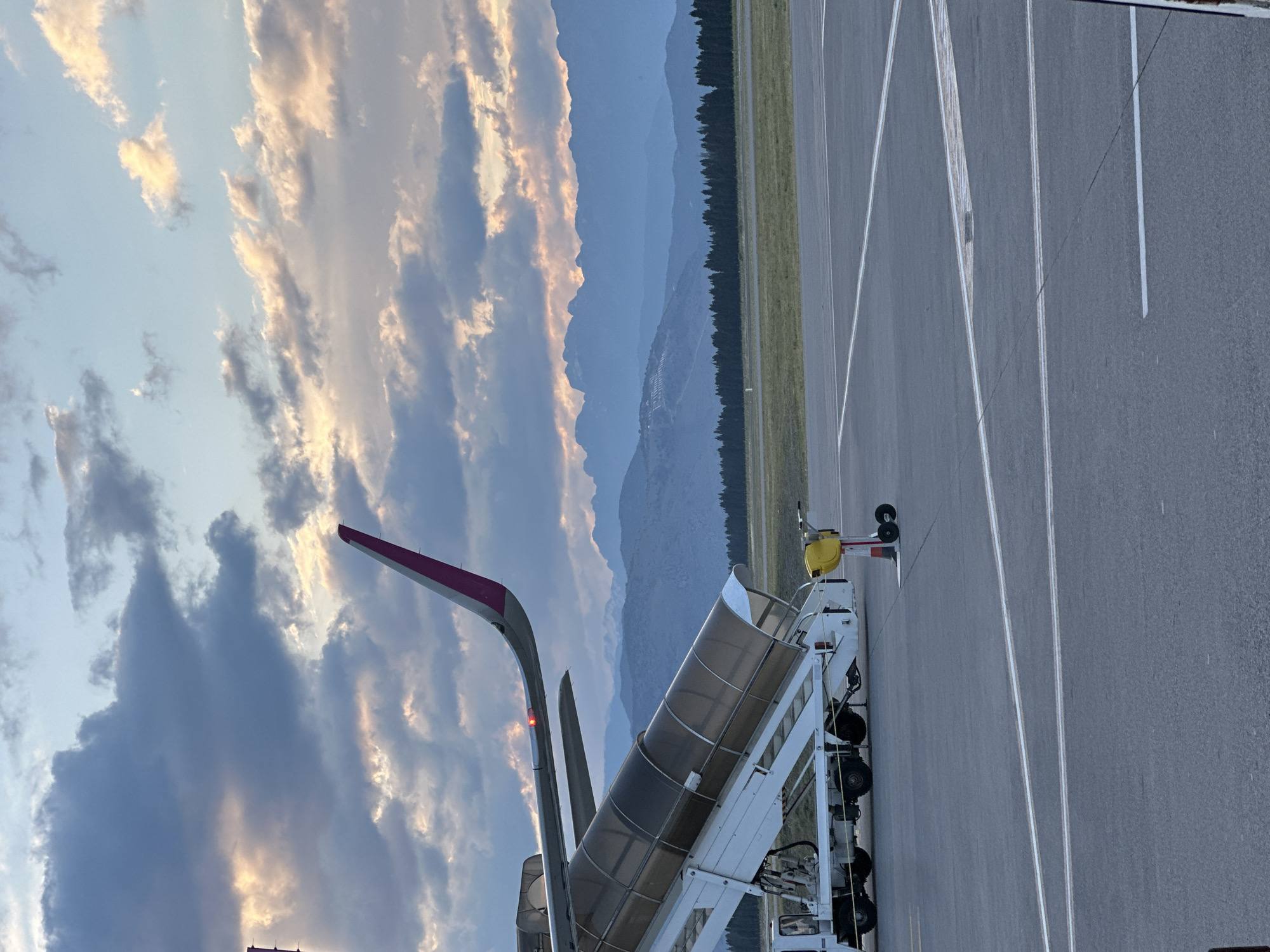 The image shows an airplane parked at an airport in Donja Cijevna, Montenegro. The airplane is connected to a mobile stairway, presumably for passengers to deboard. In the background, a mountain range is visible under a cloudy sky. The airport tarmac is marked with white lines. There are no people visible in the image. The scene appears to be at dusk or dawn, based on the lighting conditions.