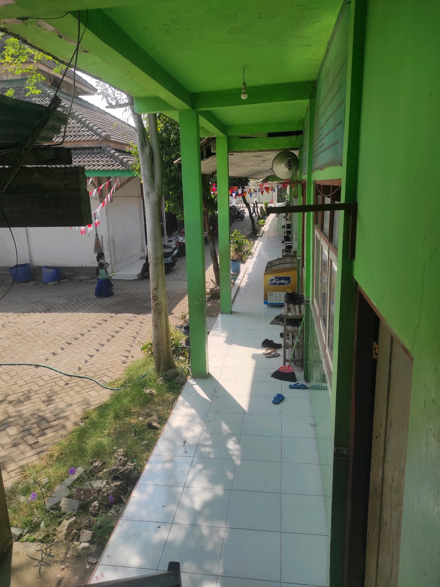 The image captures a sunlit outdoor corridor of a building with bright green painted walls and ceiling. The corridor floor is tiled with white squares. To the left of the corridor, a pathway made of beige interlocking bricks is visible, with patches of green grass and some purple flowers growing along the edge. A tall, light-barked tree stands in the middle ground. In the distance, on the brick pathway, two young children are seen, one wearing a blue skirt and a light-colored shirt, and the other in a darker outfit. They appear to be standing near a white building with a tiled roof, adorned with small red and white triangular flags. Several blue plastic barrels are also present on the pathway. Further down the corridor, a yellow freezer with the brand name "Joyday" is visible, along with a broom and several pairs of sandals on the floor. The environment suggests a school or a similar public institution in Klepek, Indonesia, given the presence of children, the flags, and the architecture. The lighting indicates it is daytime, with strong shadows cast by the overhead structures and the tree.