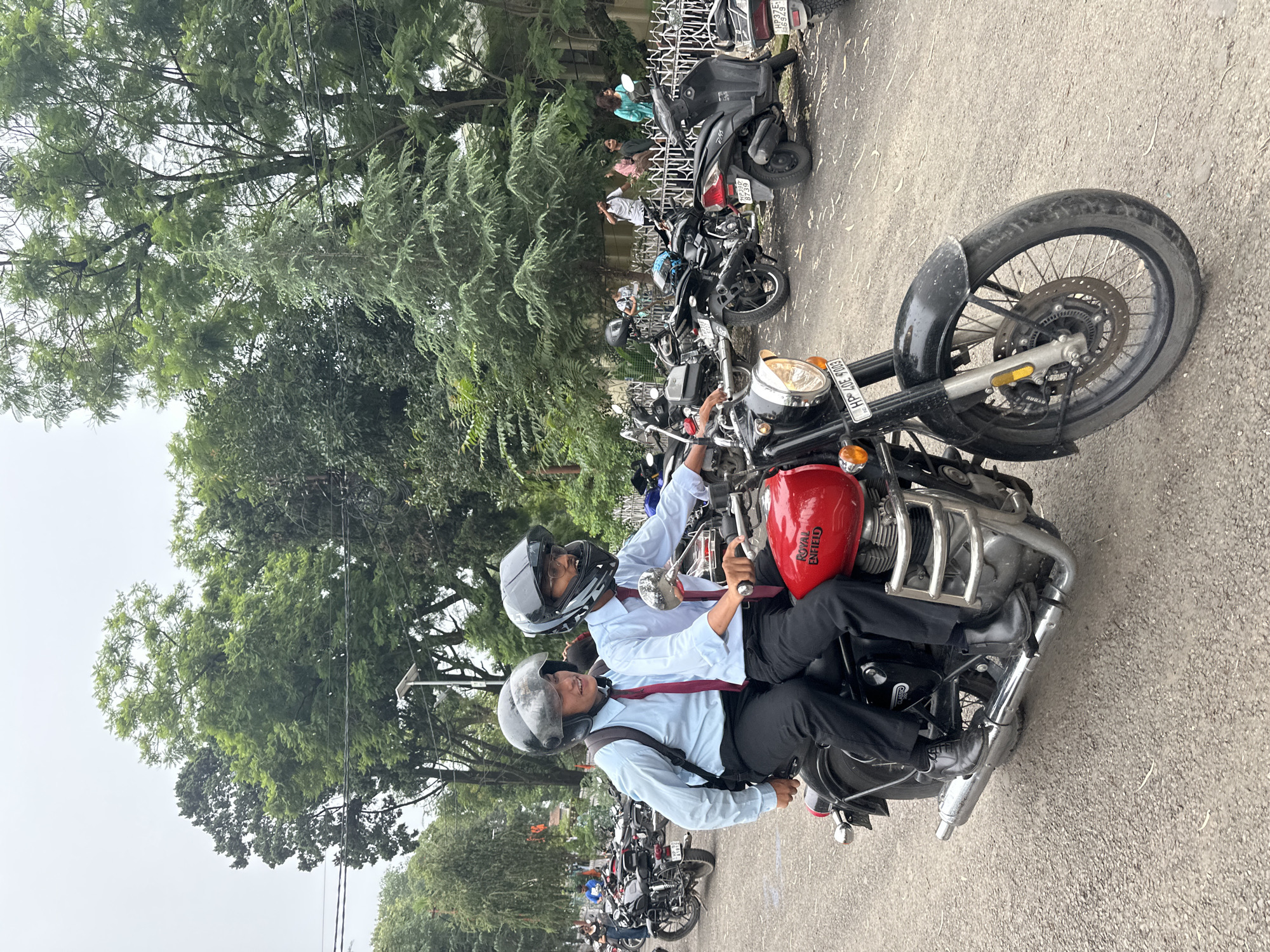 Two young people wearing light blue shirts, red ties, black pants, and helmets are riding on a red Royal Enfield motorcycle. The person in front is driving, and the person behind them is a passenger. Both individuals appear to be wearing backpacks. The motorcycle is a classic model with a chrome exhaust pipe and front crash guard. In the background, there are several other motorcycles and scooters parked. Trees and foliage are visible, suggesting an outdoor location, possibly a street or parking area in a city. The sky is overcast.
