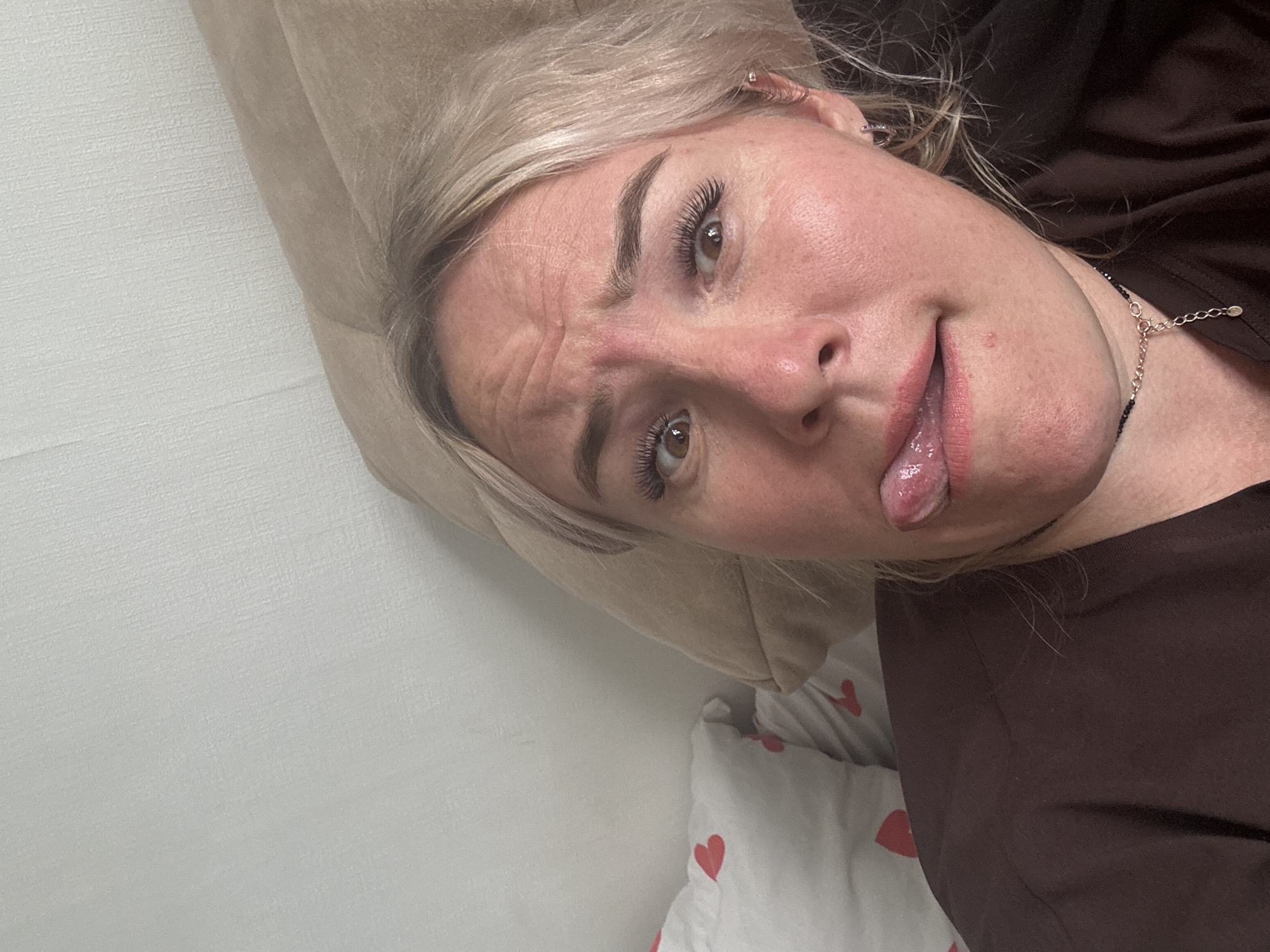 This is a selfie of a blonde woman in a dark brown top, sticking her tongue out. She has on false eyelashes and a necklace. The background appears to be a bed with a pillow and a white duvet with red hearts. The image was taken in Pozdyshki, Russia.