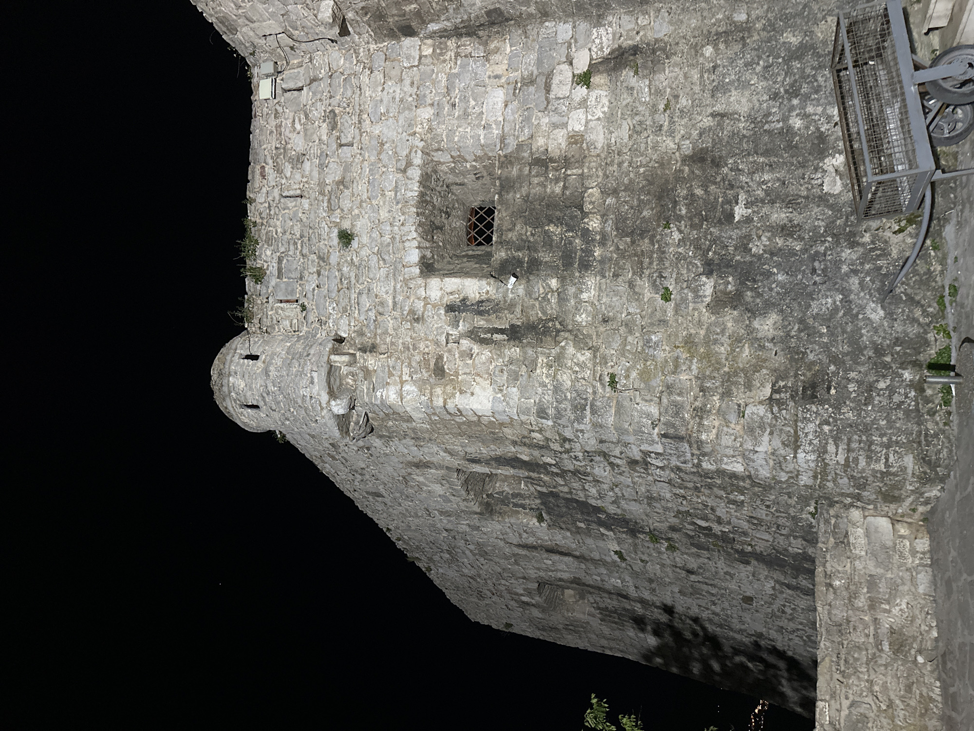 The image shows a tall, stone structure, likely a part of the city walls of Budva, Montenegro. The tower is constructed of gray stone blocks, and is weathered. The structure has a small window with a metal grate. To the right of the tower, there is a metallic cart with wheels. The background is dark, suggesting the photo was taken at night. A portion of the city wall can be seen on the right edge of the image.