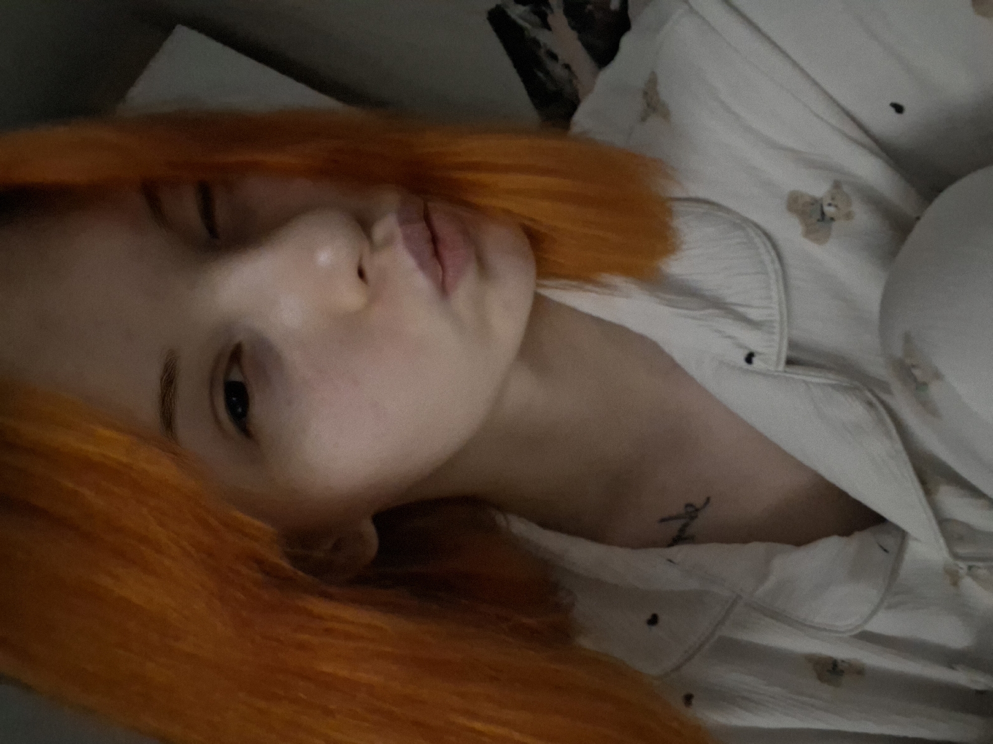 This close-up, diagonal shot features a person with striking, vibrant orange hair, lying down in what appears to be an indoor setting, likely in Chaikovsky, Russia. The individual has fair skin and is wearing a light-colored pajama top patterned with small, light brown teddy bears and tiny dark dots.
The person's face is central to the image, with one eye winking or partially closed, and the other eye visible looking towards the viewer. Their lips are slightly parted in a gentle pout. A small, dark tattoo, consisting of an illegible cursive word, is visible on their upper chest or collarbone area. The background is dark and out of focus, suggesting soft or low lighting conditions, characteristic of a bedroom or relaxed evening setting. The overall mood conveyed is one of relaxation, playfulness, or casual posing for a photograph.