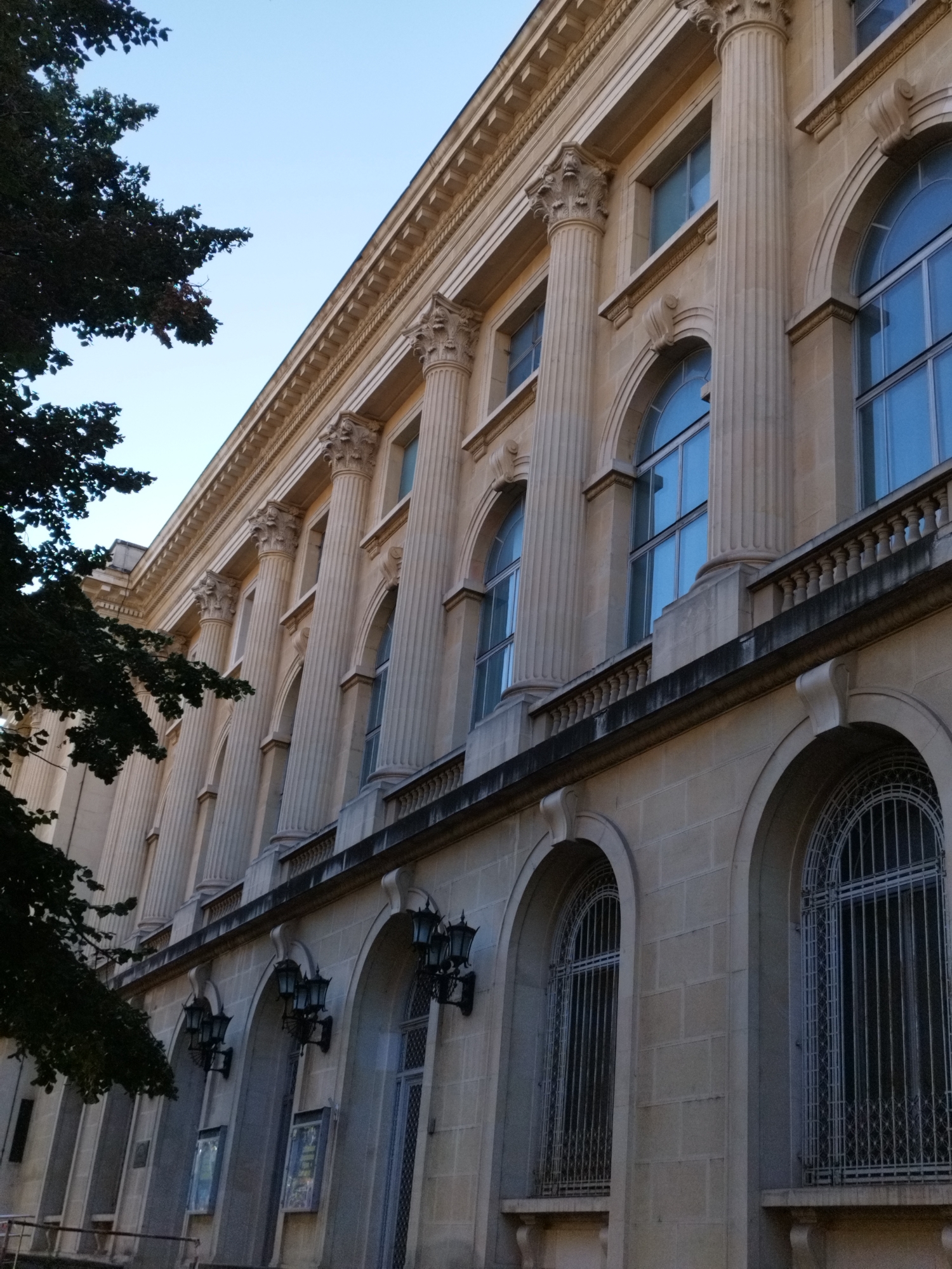 The image shows the facade of a building in Bucharest, Romania, characterized by classical architecture. The building features a series of tall, fluted columns with ornate capitals that support an entablature. Above the columns are several arched windows, some with intricate metal grilles. The facade is constructed from light-colored stone blocks. A dark green tree with dense foliage partially obscures the left side of the building and the sky. The sky above is a clear, pale blue. The lower part of the facade has arched entrances or windows with decorative grilles and ornate wall-mounted lamps. The overall impression is of an imposing and grand edifice, typical of public or institutional buildings from the late 19th or early 20th century.