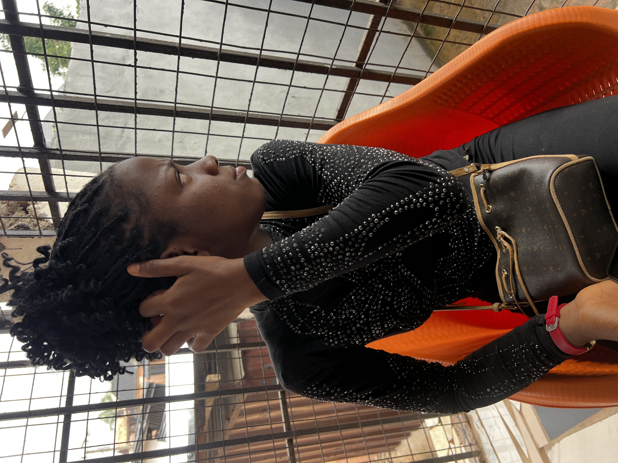A young Black woman with dark, curly hair styled in braids is seated in a bright orange plastic chair. She is wearing a long-sleeved black shirt adorned with numerous small, reflective studs. A brown, monogrammed drawstring bag with leather trim is slung across her body. A pink bracelet is visible on her left wrist. She is looking away from the camera, her left hand resting on the back of her head. The scene is framed by a rusty metal grate, suggesting she is indoors looking out through a window or bars. In the background, there are glimpses of buildings and trees, consistent with an urban or semi-urban environment in Nkpor, Nigeria. The lighting appears to be natural daylight.