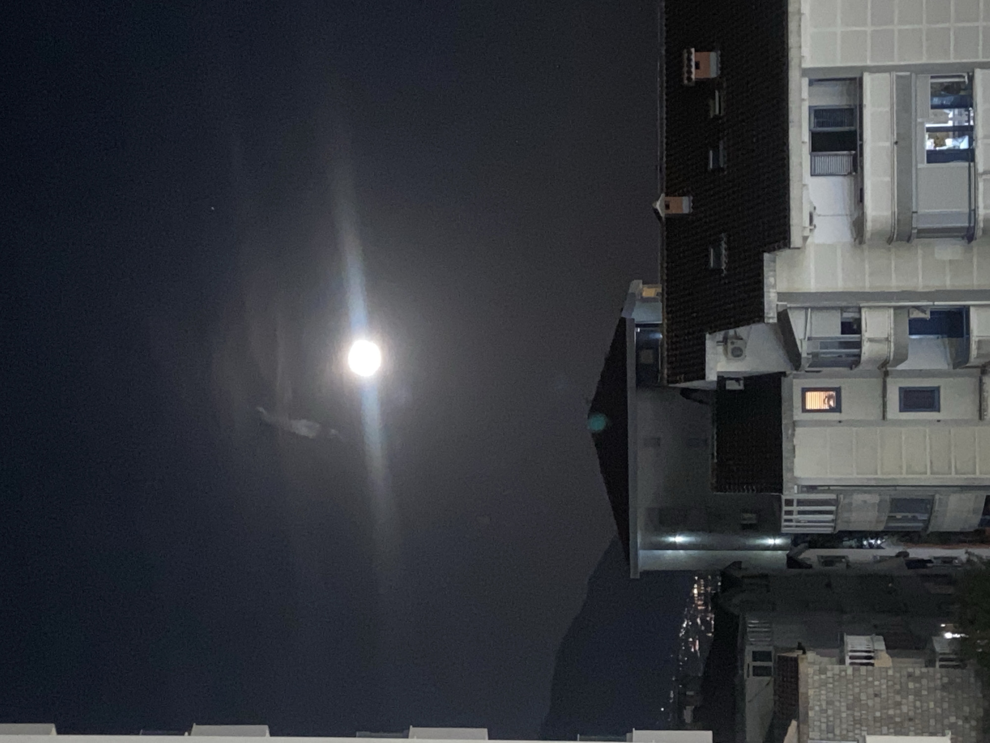 The image depicts an urban nighttime scene. A bright moon dominates the left side of the image, illuminating the dark sky. On the right, a multi-story building with white and dark gray sections stands, featuring windows, some of which are lit. Below the building, there is a cityscape with streetlights. The location is likely in Budva, Montenegro, given the building architecture and general urban environment. There are no discernible people or explicit actions visible. The image primarily showcases the contrast between the natural moonlight and the artificial light of the city.