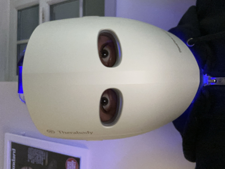 A white Therabody facial mask is shown, with two oval cutouts revealing the eyes of the wearer. The mask is held in place by a blue light strap. Part of the wearer's dark clothing is visible on the right. The background includes a framed picture on the wall and a window with white trim. The image is somewhat blurry and taken at an angle.