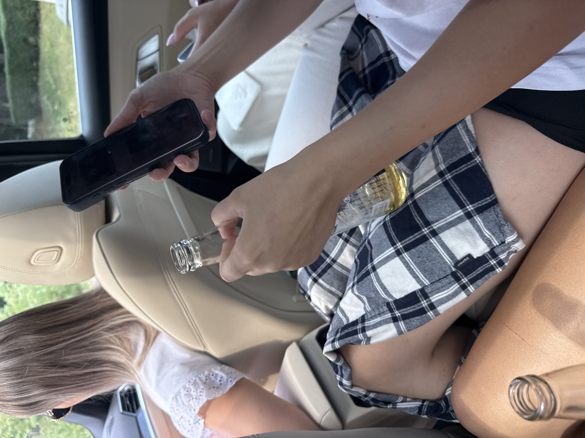 The image depicts the interior of a vehicle, likely in a rural or suburban setting given the glimpse of greenery and trees visible through the window. Two individuals are present. The person in the foreground is seated, wearing a black and white plaid skirt and a white t-shirt. They are holding a glass bottle with a yellow liquid, likely beer, in their right hand and a cell phone in their left. Another person, partially visible in the background, is wearing a white top and is seated in the driver's seat. The car's interior appears to be a light beige color, with leather seats. The scene is illuminated by natural light. The location is identified as Rusca, Moldova.