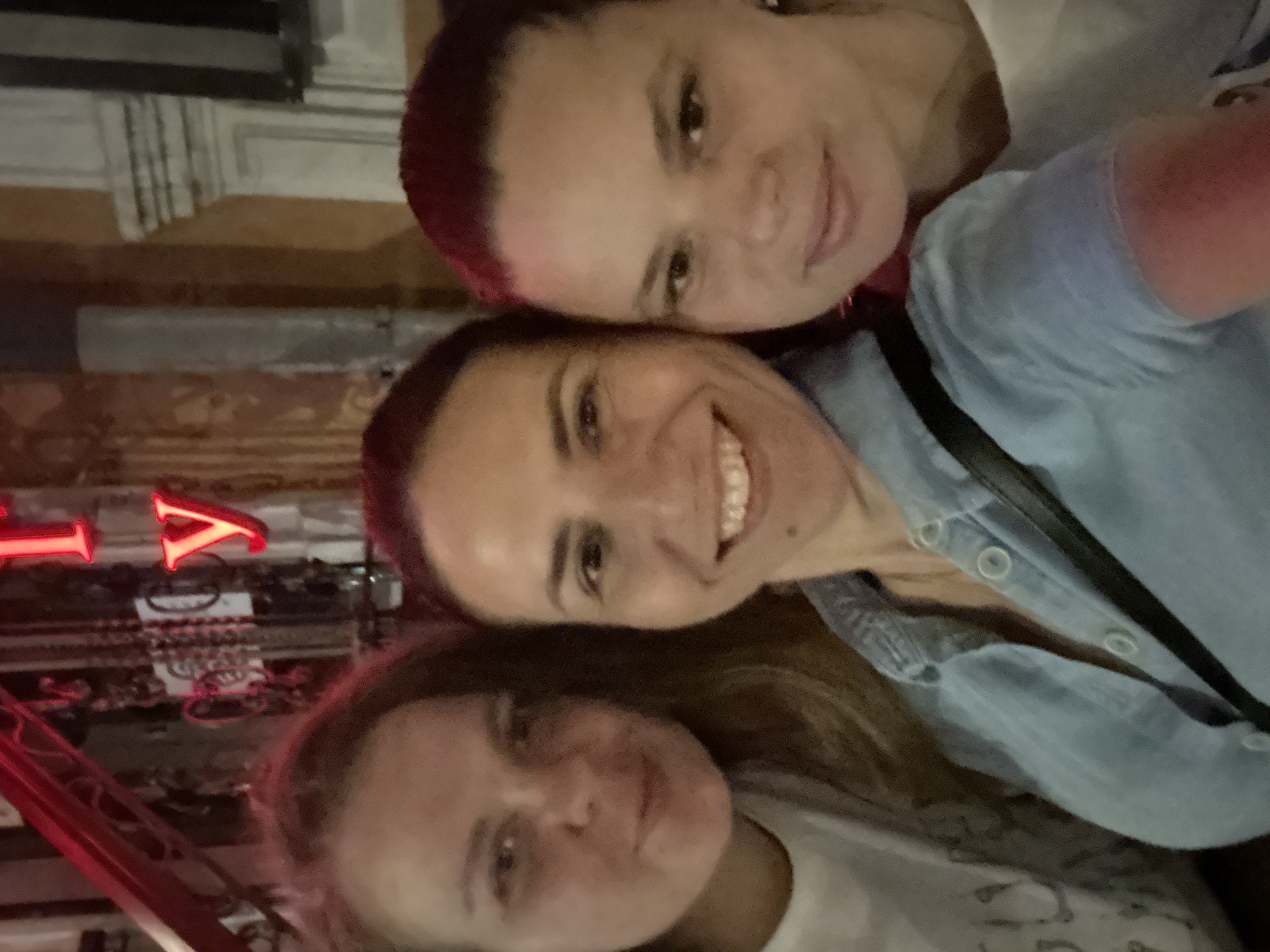 Three women are posing for a selfie outdoors at night. The woman in the center is wearing a light blue denim shirt, and the woman on the right has dark red hair. Behind them, neon signs with Cyrillic letters, one of which appears to spell "ry," are visible. The background also shows architectural details characteristic of Odesa, Ukraine, including ornate stucco work and columns. The lighting suggests a vibrant urban nightlife setting.