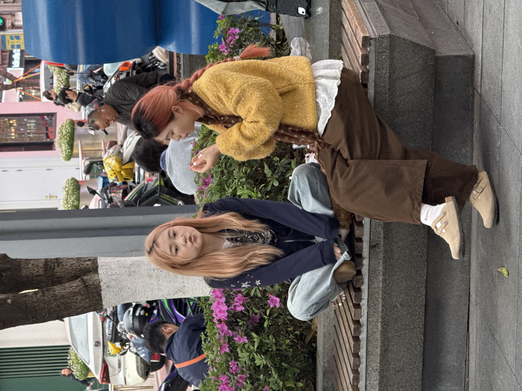 Two young women sit on a concrete bench surrounded by pink flowering bushes in what appears to be a city street setting. One woman has reddish-pink hair styled in braids, wearing a yellow textured sweater, brown pants, and tan shoes with visible white socks. She is leaning forward, looking at something in her hands, possibly flowers. The second woman has long, light-colored hair, wearing a dark blue jacket over a light-colored outfit and light blue jeans. Her knees are bent, and she faces the camera. In the background, there are other people, motorbikes, cars, and greenery. The setting is Liangjiang, China.