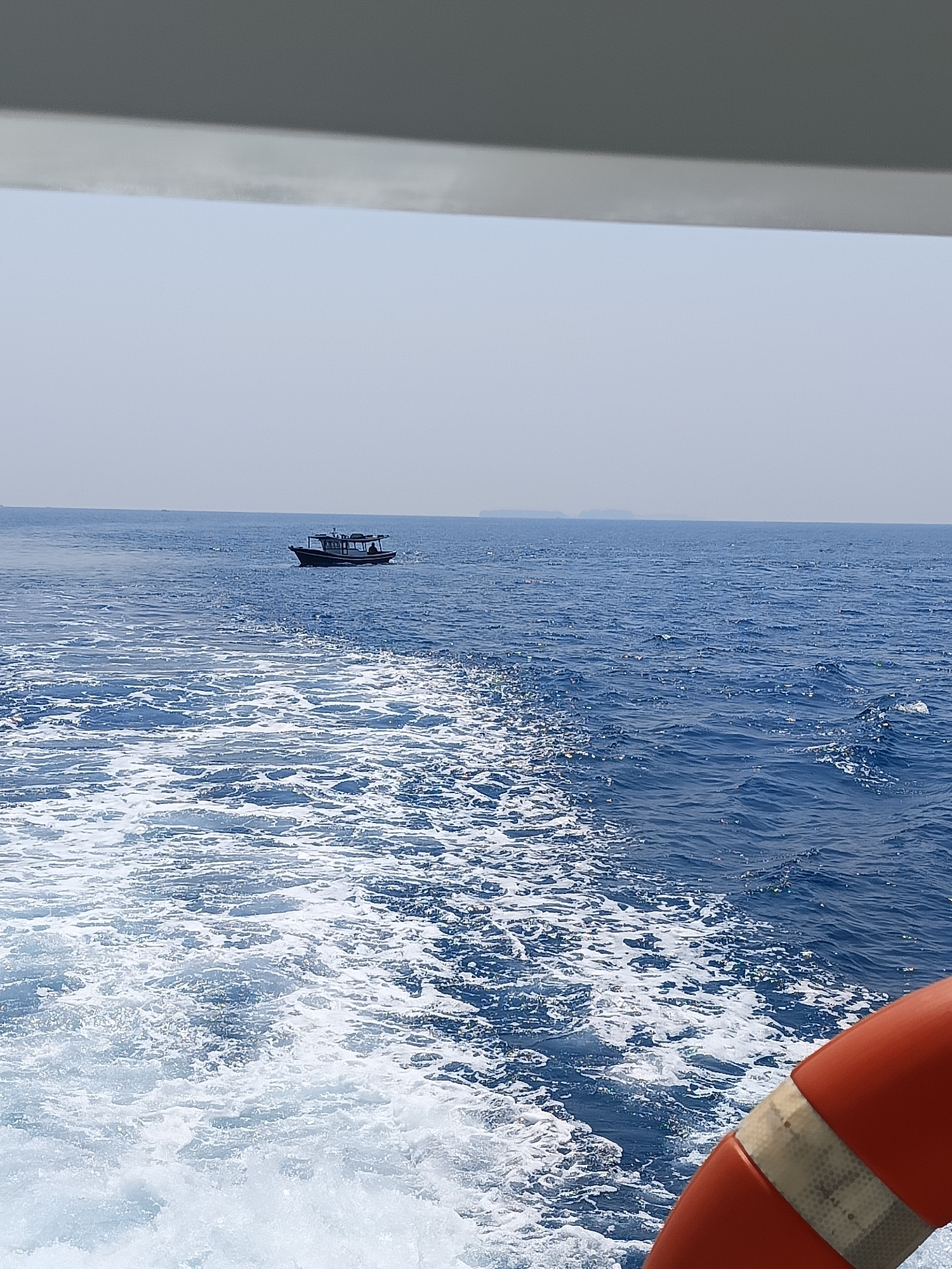 The image captures a view from a boat on the sea, looking towards a distant boat. The foreground shows the white wake of the current boat, with foamy water extending towards the middle ground. In the middle ground, a small, dark-colored boat with a roof and at least two visible people on board is moving across the blue water. The horizon is visible in the background, with a hazy sky above. A glimpse of an orange life preserver is visible in the lower right corner of the frame. The scene is characteristic of the waters surrounding the Special Capital Region of Jakarta, Indonesia, possibly in the Thousand Islands archipelago.