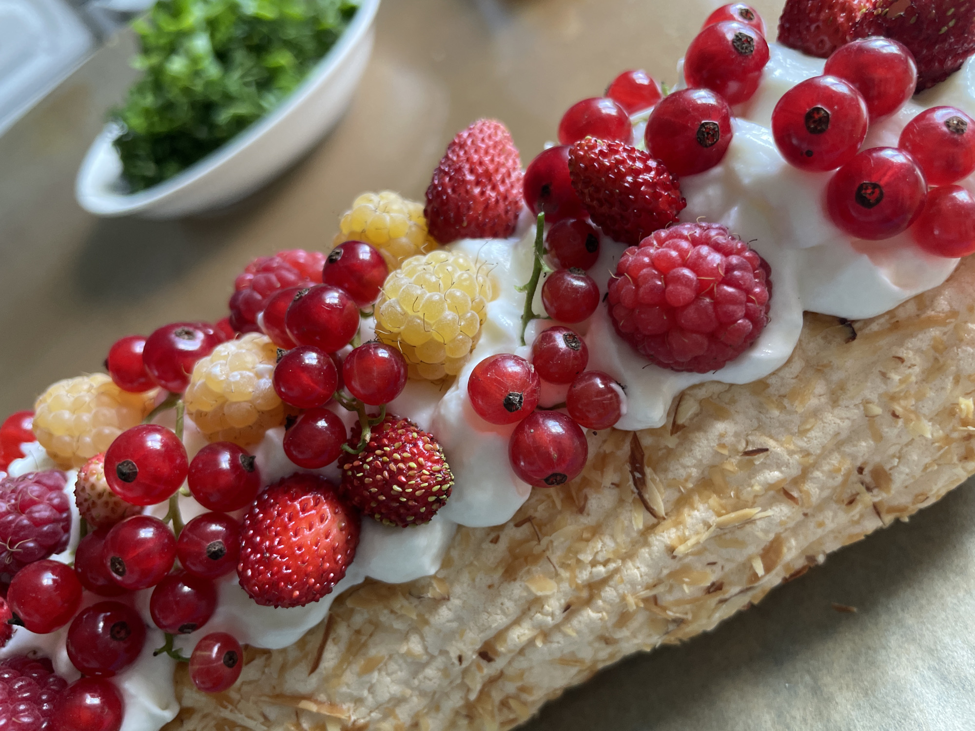 A close-up, high-angle shot shows a portion of a meringue dessert topped with whipped cream and various berries, including raspberries, red currants, and wild strawberries. Some of the berries are still attached to their stems. The meringue itself appears textured and coated with slivered almonds. In the background, a white bowl filled with chopped green herbs is visible on a light brown surface. There are no people present in the image, and no specific location in Дресвяново, Russia, can be identified.