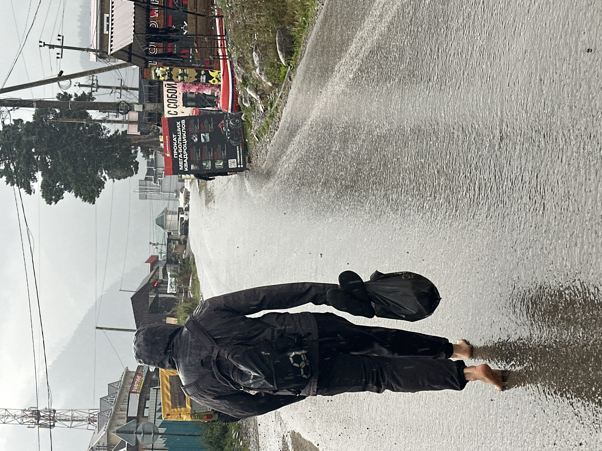 In Arkhyz, Russia, on a rainy day, a person walks barefoot along a wet, paved road. They are wearing dark clothing, including a hooded jacket and pants, and a backpack. In their right hand, they carry a black plastic bag. The road is reflective with puddles and ripples from the rain, and small droplets are visible in the air. Buildings and shops line the right side of the road, some with awnings. Signs are visible, including one advertising quad bike rentals. To the left, there are more buildings, a yellow vehicle, and a tall telecommunications tower. Mountains are visible in the background under a cloudy sky.