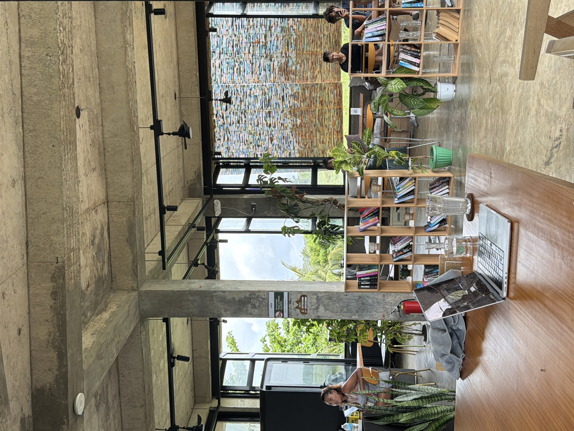 The image captures the interior of a cafe in General Luna, Philippines. The location features an industrial-style design with exposed concrete beams and black metal framework. A seating area is arranged near large windows, providing natural light and views of green foliage outside. Two individuals are seen seated in the background. Bookshelves with books and plants are set along the wall. A wooden table is the focus, with a laptop, glasses, and a folded gray shirt placed on top. A person is standing at a counter with plants nearby.