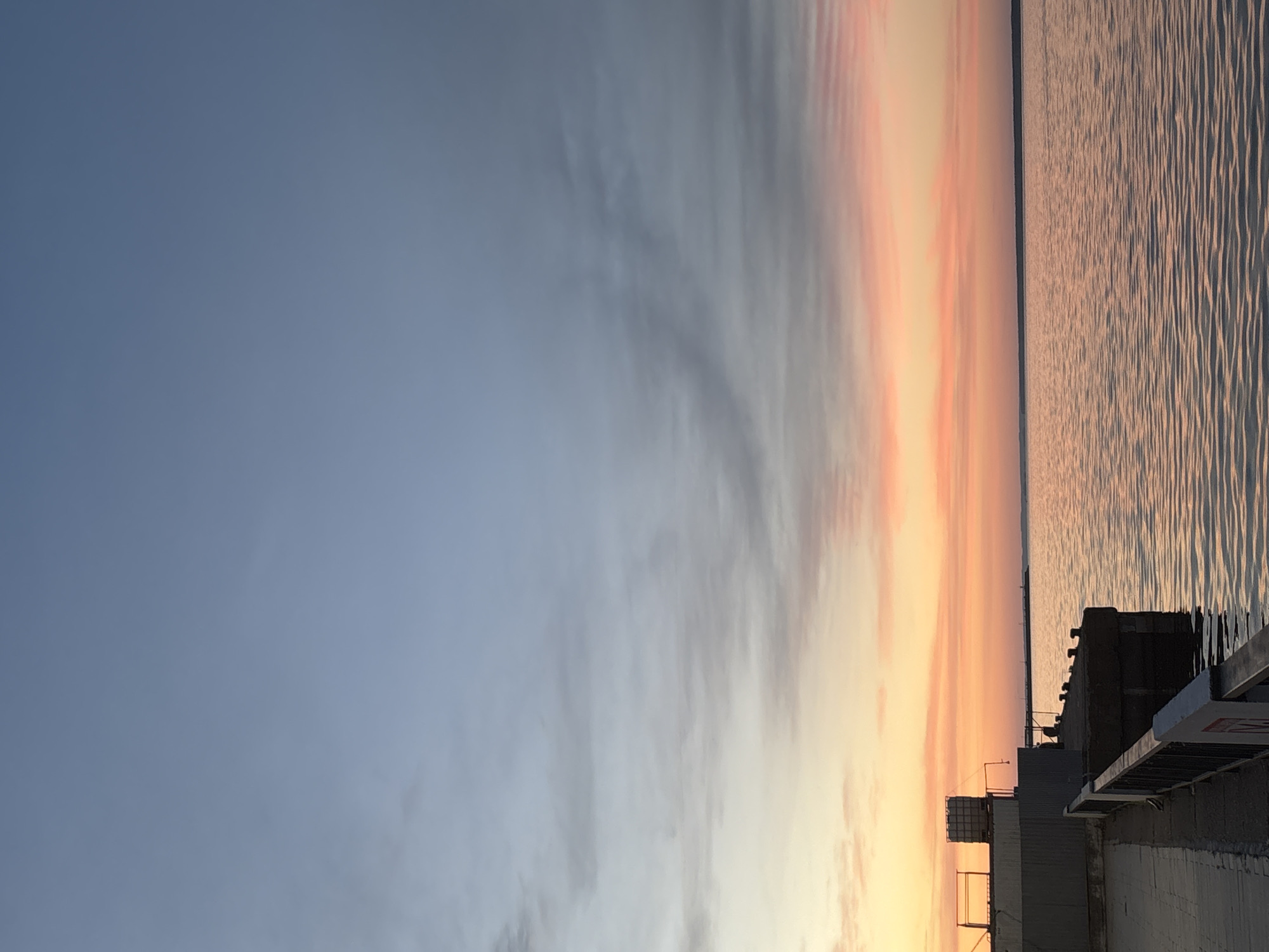 An image captures a serene waterfront scene in Chaikovsky, Russia, during a soft sunset. The sky transitions from a pale blue at the top to hues of pink and orange near the horizon, with wispy clouds scattered across the expanse. The calm waters of the Kama River reflect the warm colors of the sky, creating a shimmering effect on the surface.
In the foreground, a concrete pier extends into the water, with a metal railing running along its edge. Some industrial structures, including what appear to be large containers or storage units, are visible on the left side of the pier. The architecture suggests a working port or industrial area adjacent to the river.
The overall atmosphere is tranquil, with the fading light casting a peaceful glow over the landscape. No people are visible in the image. The scene depicts a quiet moment by the river, highlighting the natural beauty of the sky and water contrasted with the man-made structures of the port.