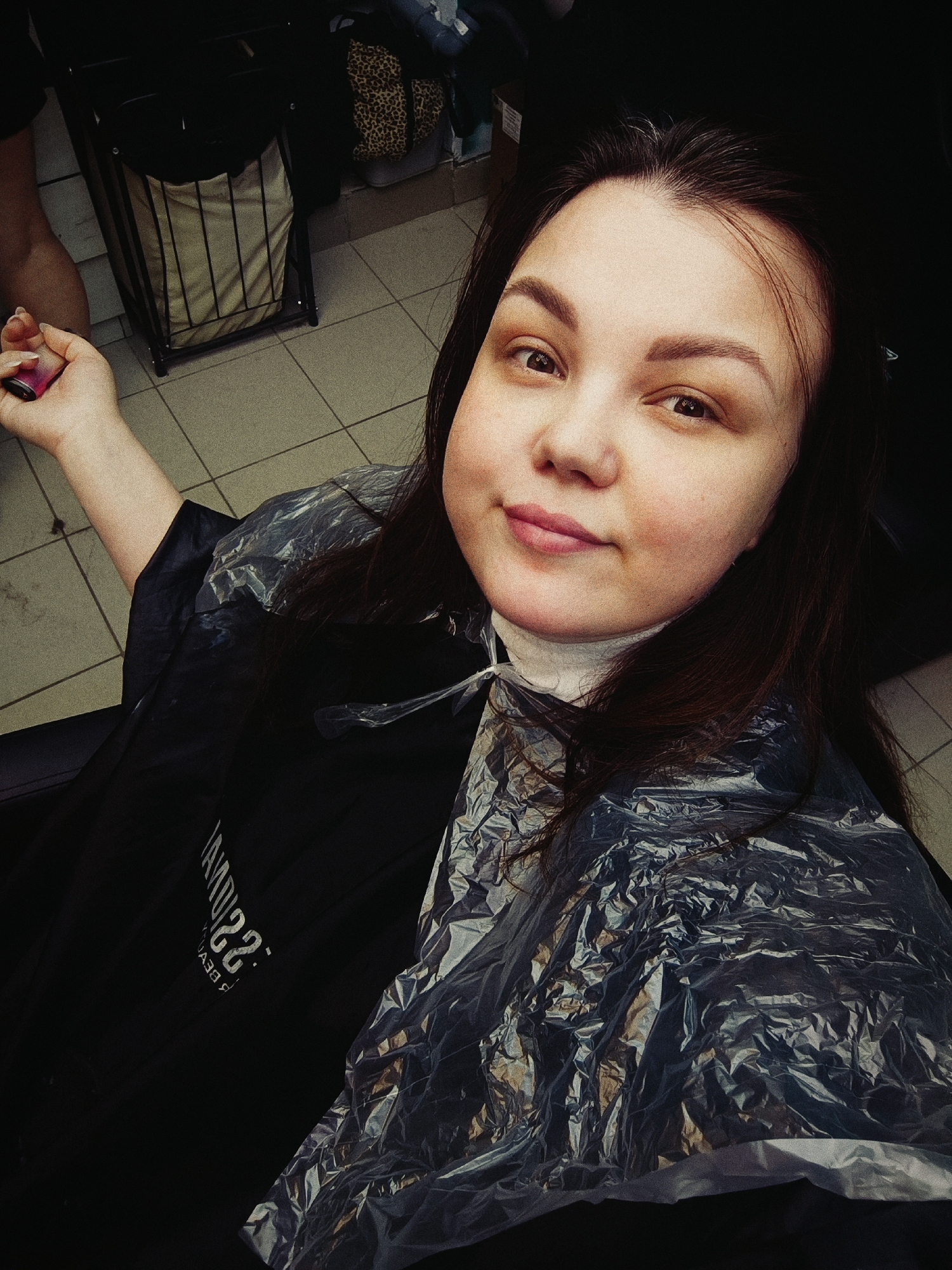 The image shows a woman with long brown hair and fair skin, wearing a plastic salon cape, possibly in a hair salon in the city of Unknown, Unknown. The cape has a crinkled, reflective texture, and a black underlayer. In the background, there's a basket and what appears to be a tiled floor. She is holding something in her hand.