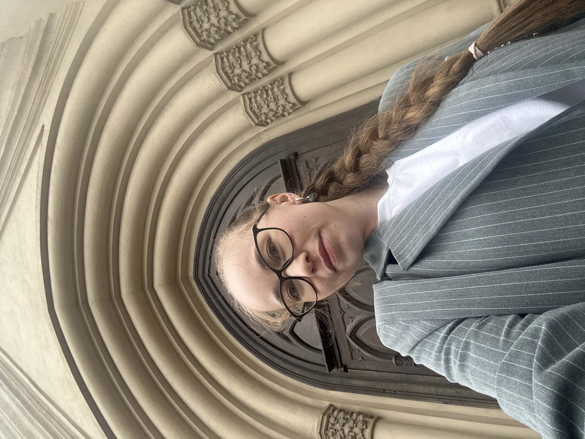A woman stands in front of a large arched doorway. She has blonde hair, which is styled in a braid. She is wearing glasses, a white shirt, and a gray pinstripe blazer. The doorway has a multi-layered arched design with decorative elements. The doorway itself is dark wood with carved details. The location is the archway of St. Isaac's Cathedral in Saint Petersburg, Russia.