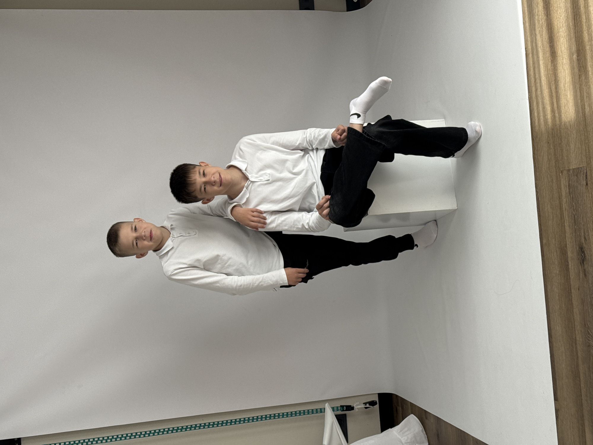 Two boys are posing for a photo in a studio with a white backdrop and a light wood-look floor. The boy on the left stands while the boy on the right sits on a white cube. Both boys are wearing white shirts and black pants, with white socks. The boy on the right has a Nike logo visible on one sock. The boy on the left has his arm around the boy on the right. The scene appears to be a professional photoshoot.
There is no information in the image to determine if this photo was taken in Izhevsk, Russia.