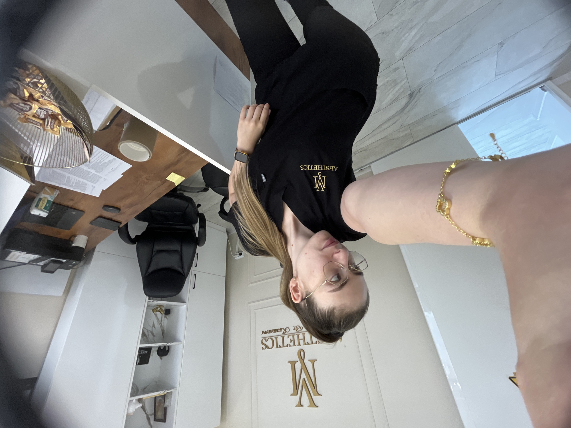 A woman in black scrubs, wearing glasses and a gold bracelet, stands in a room. She is taking a selfie. The room appears to be a medical office or clinic, with white walls, a desk, and a shelf with decorative items. There is a logo on the wall behind her that reads "M AESTHETICS Dr. Kuzmanova." On the desk, there is a printer, a coffee mug, and a bowl of candy.