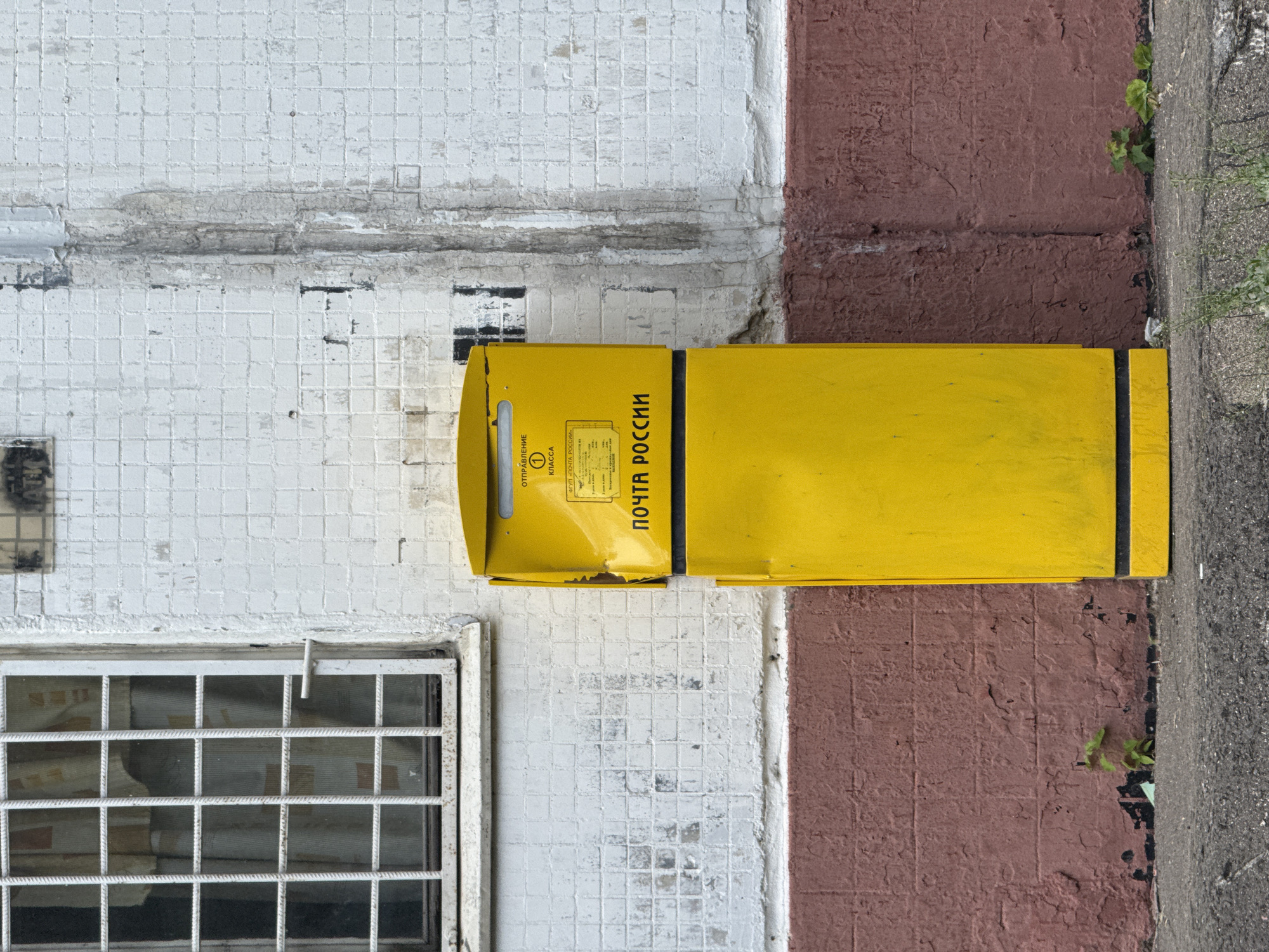 A yellow, rectangular postal box stands against a white, tiled building. The postal box has a flap at the top for inserting mail, and text in Russian characters is visible on its surface. The building has a grimy, weathered appearance, with vertical streaks of discoloration running down the wall. To the left of the postal box is a barred window, behind which a light-colored curtain is visible. The lower portion of the building is painted a dark red color. In front of the building, a grey asphalt sidewalk is visible, with small plants growing near the base of the wall. The scene suggests an urban environment, likely in Russia, given the Cyrillic text on the postal box.