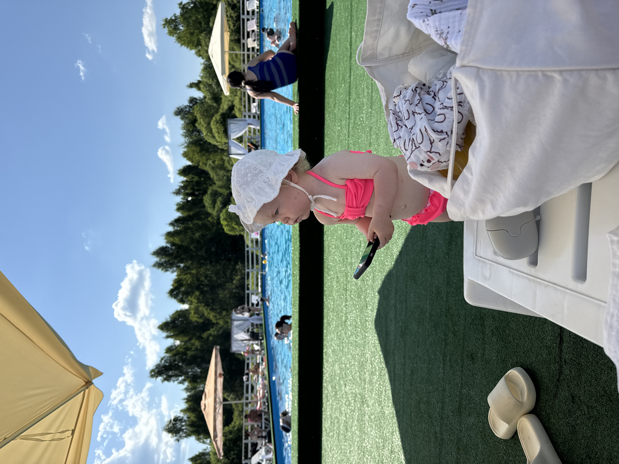A young child wearing a white sun hat and a pink bikini is the main subject of this image, which appears to have been taken in Koloksha, Russia. The child is looking down and holding a black object, possibly a toy or a phone. The setting is an outdoor swimming pool area on a sunny day, indicated by the bright blue sky and white clouds.
In the background, other people are visible in and around the pool. Some are swimming, while others are relaxing. There are lounge chairs and umbrellas scattered around the poolside, and a bright green, artificial turf covers the ground. The image is oriented vertically, with the sky taking up a significant portion of the left side. The overall mood seems relaxed and summery, consistent with a day at a pool. There is no visible text in the image.