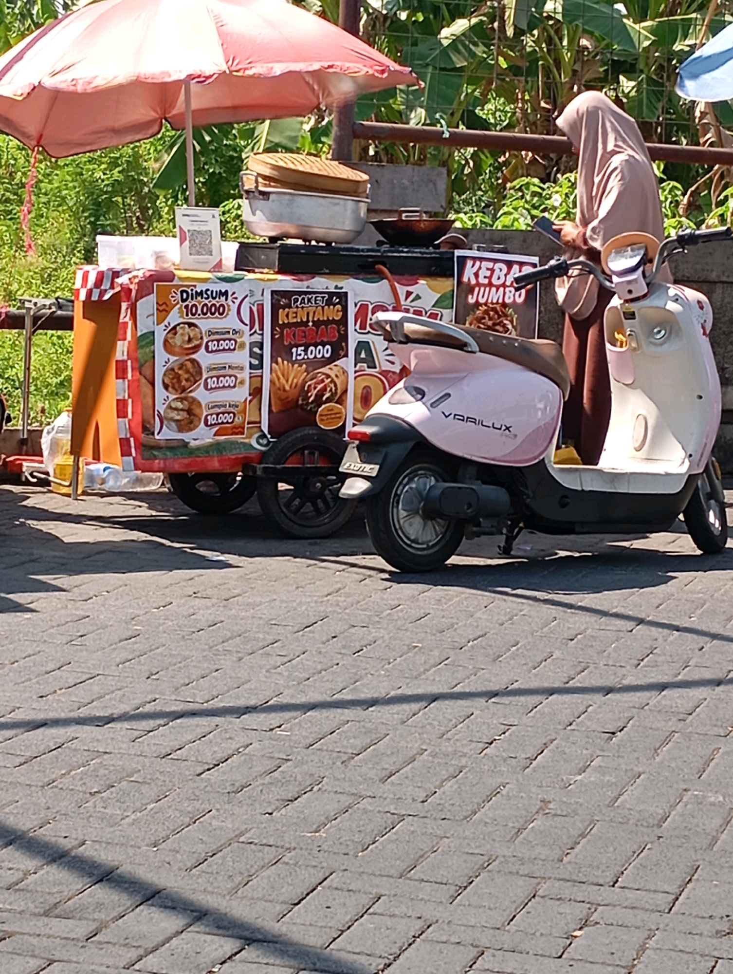 In Boro, Indonesia, a food cart is parked on a brick sidewalk. The cart displays a menu with items like "Dimsum" and "Paket Kentang Kebab Jumbo," with prices listed in Indonesian Rupiah. To the right of the cart, a person wearing a light brown hijab stands next to a pink and white electric scooter. The scooter has the word "VARILUX" written on its side. In the background, lush green foliage is visible, with a partial view of a red and white striped umbrella over the food cart. The scene is bathed in sunlight, casting shadows on the pavement.