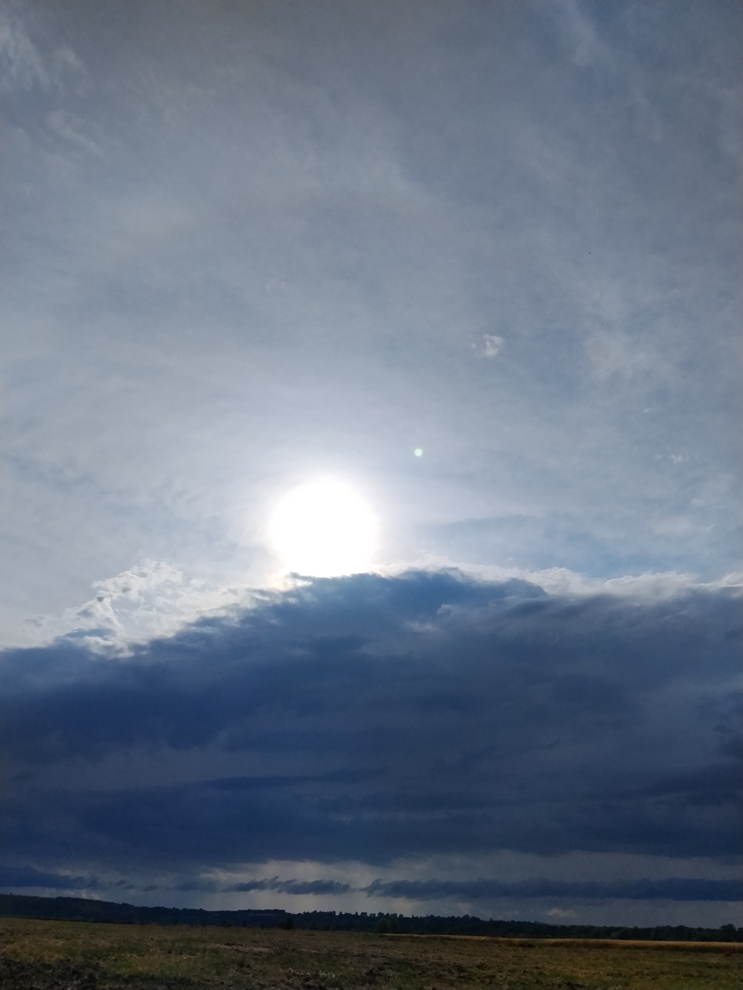 The image depicts a dramatic sky over a rural landscape. The sun is a bright, diffuse disc partially obscured by a thick band of dark, ominous clouds that occupy the lower half of the frame. Above these dark clouds, lighter, wispy clouds stretch across a pale blue sky. The foreground is a field of dry, muted-color vegetation, possibly harvested or fallow, with a dark treeline and rolling hills visible on the distant horizon. There are no people or specific urban structures visible in the image, suggesting a natural or agricultural setting. Given the location of Ustyvytsya, Ukraine, this scene likely represents a transitional weather pattern, possibly the approach of a storm, common in such a region.