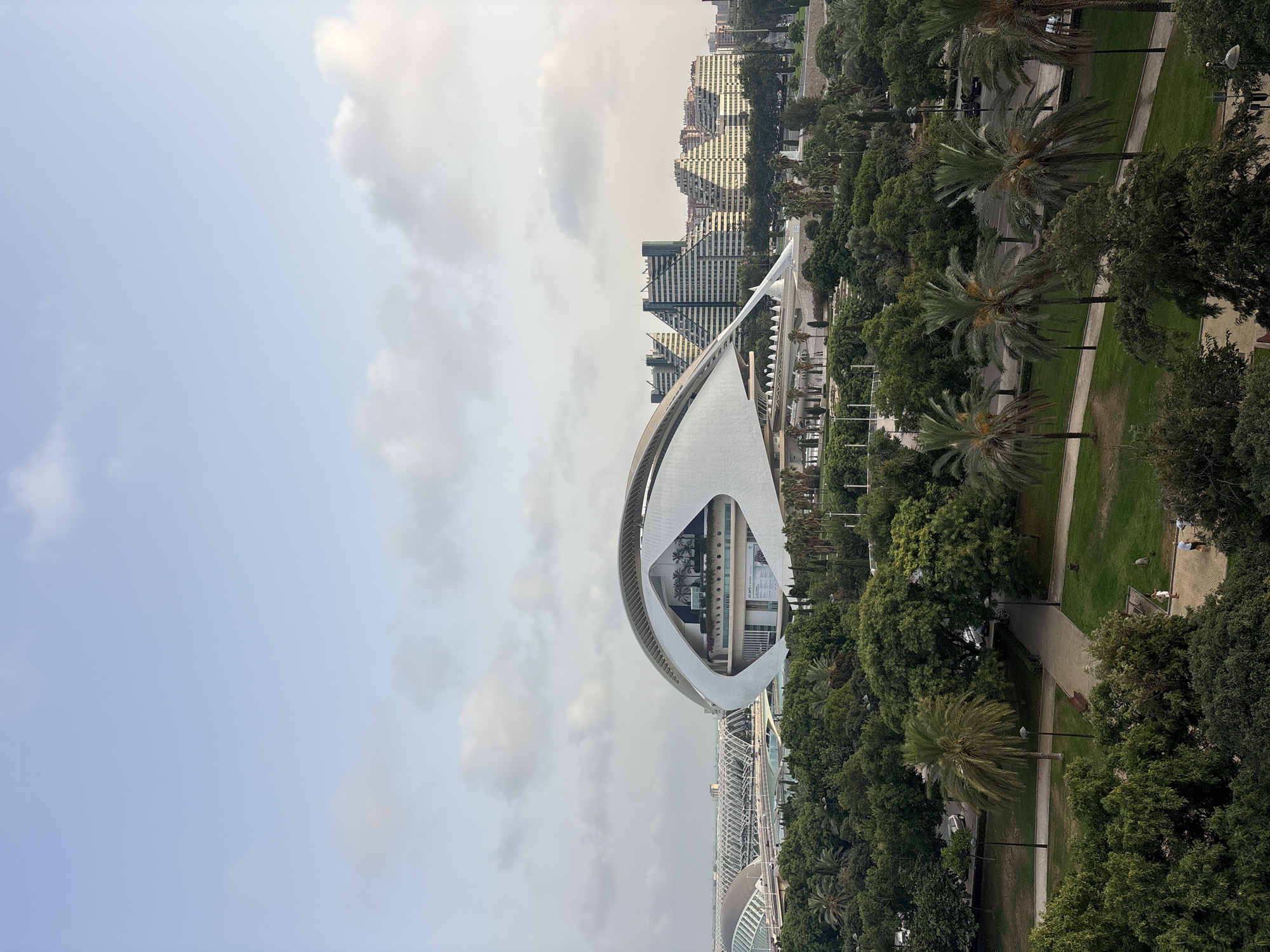 The image captures an aerial view of the City of Arts and Sciences in Valencia, Spain, under a partly cloudy sky. In the foreground, a lush green park with winding paths and scattered trees, including several palm trees, is visible. In the middle ground, the iconic Hemisfèric building, designed to resemble a human eye, stands prominently with its distinctive white, curved structure and a large opening that reveals interior elements, including palm trees on a balcony. Beyond the Hemisfèric, a series of modernist buildings with stepped, triangular facades are seen in the background, alongside other structures that appear to be part of the science complex, characterized by their white, arched roofs and bridges. Several vehicles are present on roadways within the park and in the background. A few individuals are walking in the park, one group near a paved walkway and another pair further in the distance. The overall scene is one of modern architecture integrated with verdant urban landscaping.