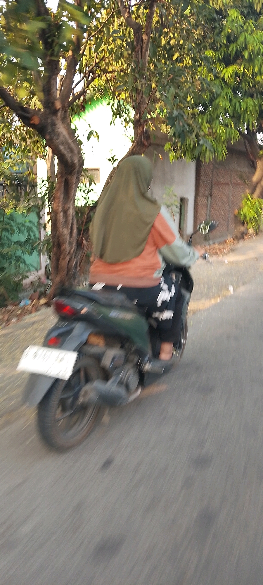 A person wearing a green hijab and an orange shirt is riding a dark-colored motorcycle on a paved road. The person is facing away from the camera and is partially obscured by the motorcycle. The motorcycle has a white license plate on the rear. The scene appears to be in a residential area with trees and buildings visible in the background. The lighting suggests it is daytime, possibly late afternoon, with sunlight filtering through the trees. The motion blur indicates the motorcycle is in motion. The location is identified as Bekasi, Indonesia.