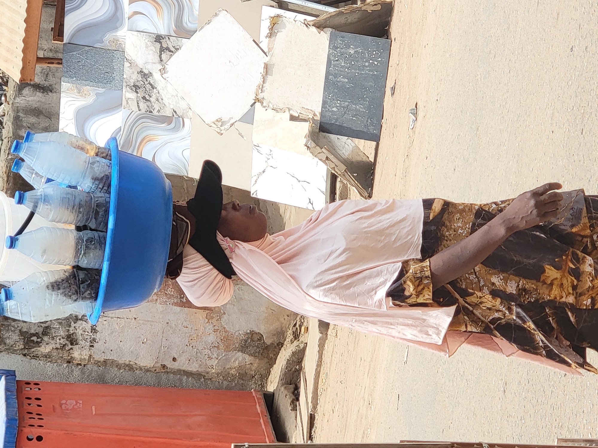 A woman carries a blue basin filled with bottled water on her head in Maiburiji, Nigeria. She is wearing a light pink top and a brown patterned skirt, with a black head covering. The background shows a mix of materials including tiles, concrete walls, and what appears to be an orange metal sheet. The ground is a sandy color. The light suggests it is daytime.
