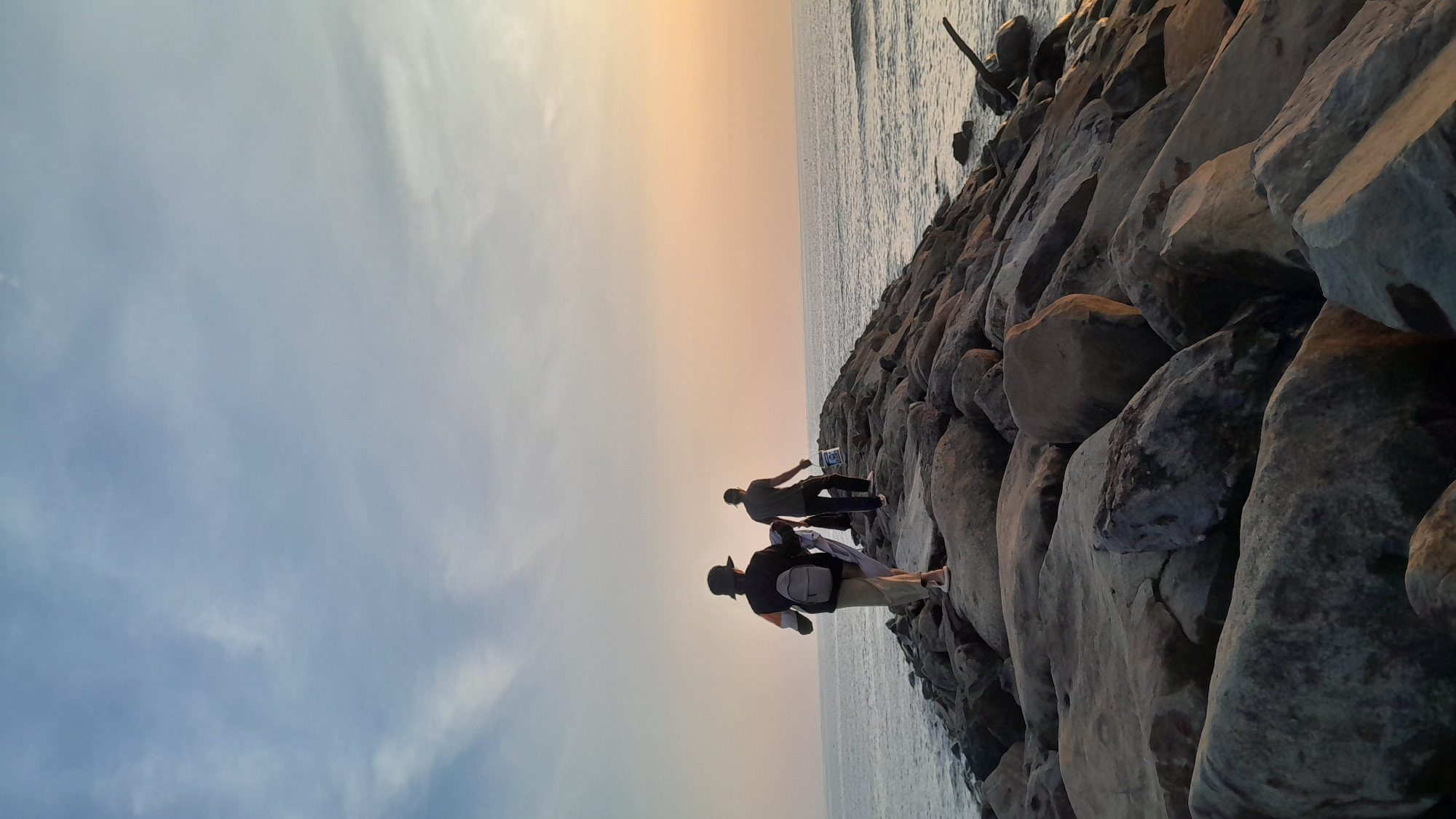 The image displays two individuals walking away from the camera along a rock jetty extending into the sea in Lhokseumawe, Indonesia. The sky transitions from light blue at the top to a warm orange-yellow hue near the horizon. The individual closer to the camera, positioned on the left side of the jetty, wears a dark top, light-colored pants, and a dark wide-brimmed hat, with a light-grey backpack on their back and a white garment draped over their left arm. The second individual, further along the jetty on the right, wears a dark short-sleeved top and dark pants, and carries a white bucket with a blue lid in their right hand. The jetty is composed of large, irregular grey rocks. The sea with visible wave activity borders both sides of the jetty. A piece of debris is partially submerged in the water on the right side of the jetty.