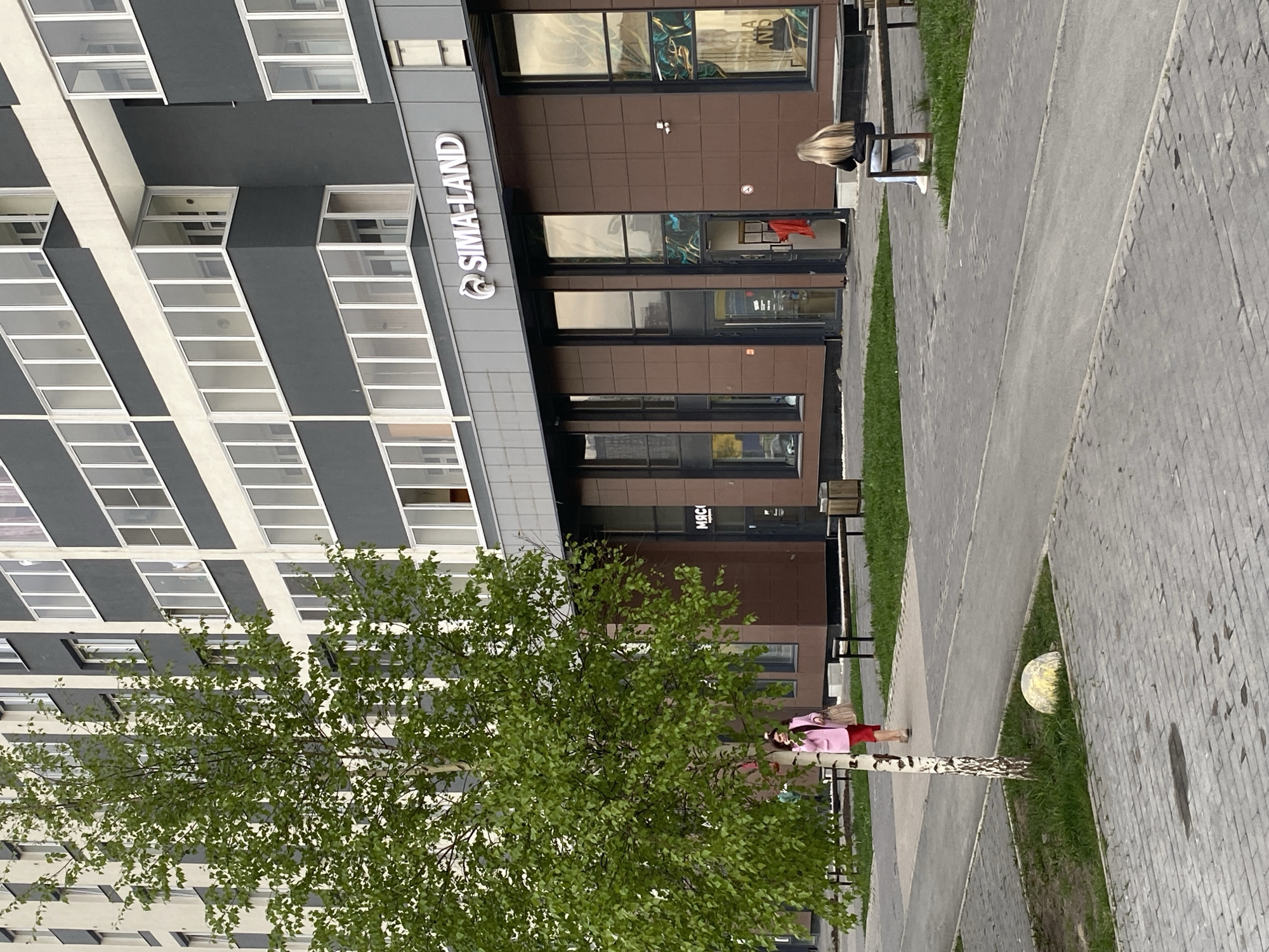 The image captures an urban scene in Yekaterinburg, Russia. A tall, modern building with alternating gray and white sections featuring rows of windows dominates the left side of the frame. The building has a brown-colored commercial section at street level, branded with "SIMA-LAND" signage.
A birch tree with lush green foliage stands in the foreground, partially obscuring the view of the building. A sidewalk runs along the right side of the building, paved with gray brick. Patches of grass are interspersed between the sidewalk and the building's facade.
A woman with long blonde hair is sitting on a bench in front of the building. A child dressed in a pink jacket and red skirt is standing near the base of the birch tree. A large, spherical object, possibly a decoration, is placed in the grassy area beside the sidewalk.