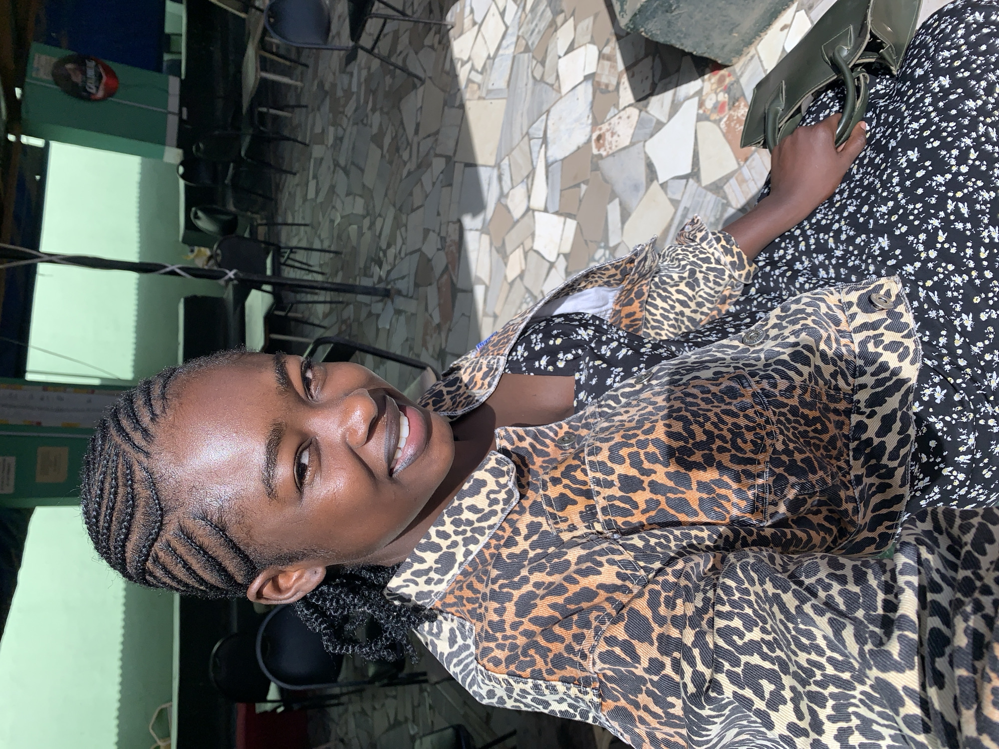 A young woman is captured smiling broadly in an outdoor setting in Kabong, Nigeria. The image is oriented sideways (rotated 90 degrees counter-clockwise). She has dark skin and her hair is styled in neat cornrows, with some braids falling to her side. She wears a black dress with a small white floral pattern, layered with a distinctive leopard print jacket. Her left hand is visible, holding a dark green handbag that rests on her lap. She looks directly towards the camera, her face lit by bright sunlight, revealing a joyful expression and teeth in her smile. A sheen on her skin suggests warmth.
The setting appears to be an outdoor patio or seating area, with a floor paved in irregular, light-colored mosaic tiles or broken concrete. Sharp shadows on the ground and background walls indicate bright sunshine, suggesting it is daytime, likely midday. In the background, green painted walls are visible, along with several dark chairs. A partially obscured red and white oval sign, possibly advertising a beverage, is visible on a green wall, with the word "Gulder" faintly legible. Some wires or lines are also strung across the scene in the background.