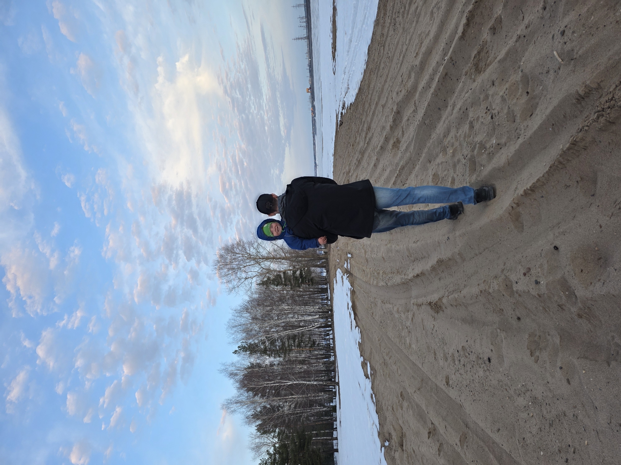 Two individuals, presumed to be an adult male and a child, stand on a sandy area. The adult is wearing a black coat and blue jeans with black shoes, and a black cap. The child has a blue jacket with a green hood. They stand near a shoreline with snow and ice. In the background, a stand of bare trees separates the sandy area from a cloudy sky. Across the expanse of water, indistinct structures resembling power lines are visible. The location is Noviy, Russia.