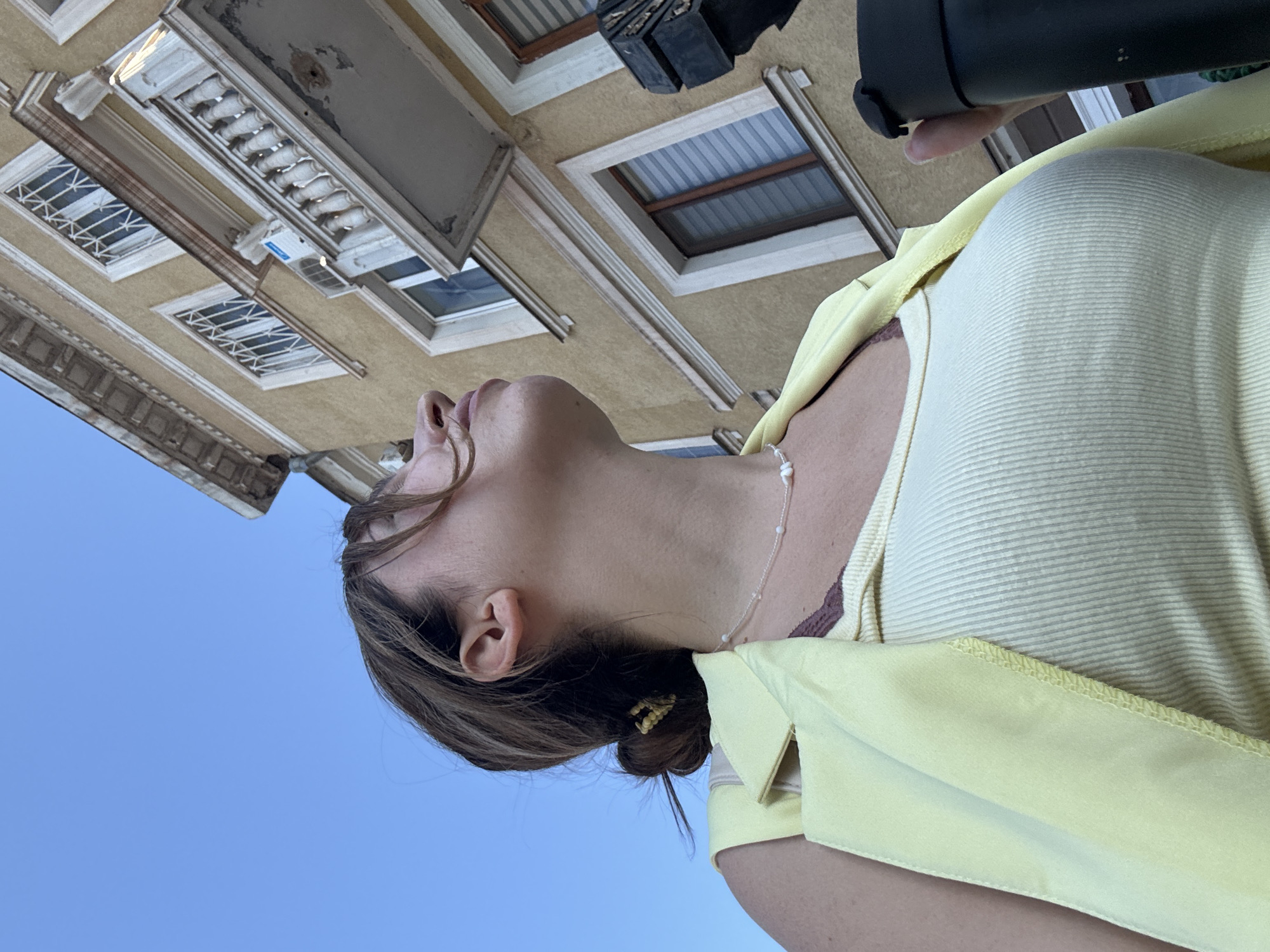 A low-angle, medium shot captures a young woman from the chest up, looking upwards. She has her hair pulled back into a bun and wears a light yellow vest over a ribbed white tank top. A beaded necklace is visible around her neck. In her right hand, she holds a black thermos. The background features a yellow building with white trim and several windows, as well as a section of blue sky. The architecture suggests a city environment, likely Kryvyi Rih, Ukraine, based on its characteristic building style. The perspective implies the photo was taken from ground level, looking up at the woman and the building.